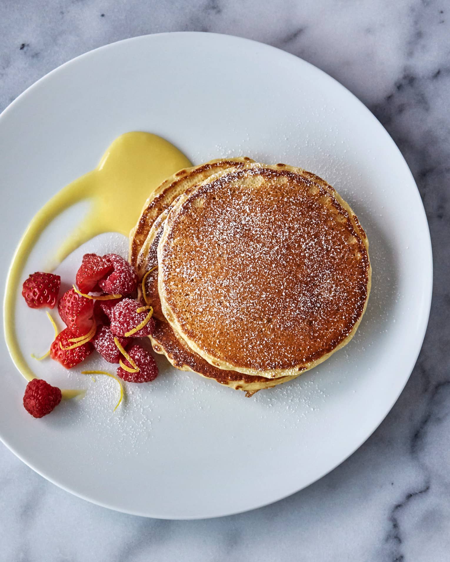 Stack of Lemon Ricotta Pancakes on plate dusted with icing sugar, lemon sauce and fresh raspberry garnish