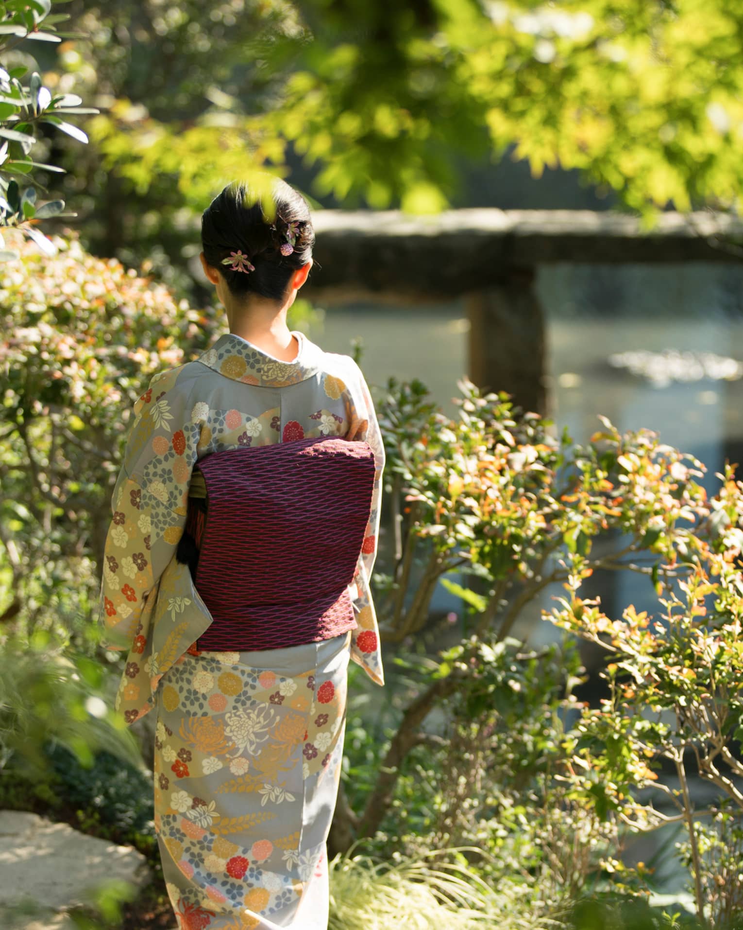 Amid bushes and trees, rear view of a person in a grey kimono with beige and orange flowers and a black and pink obi cushion.