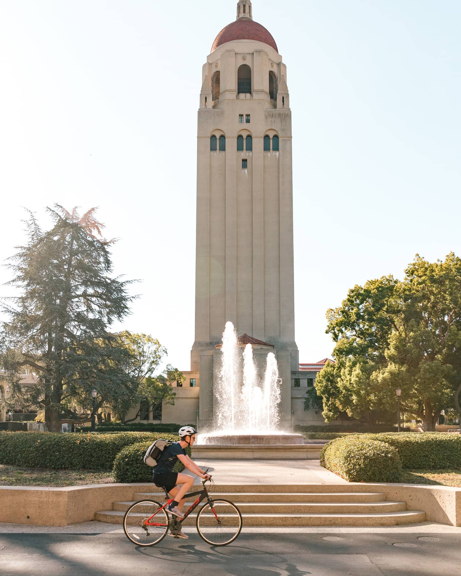 A large tower building with a biker cycling passed it in a park.