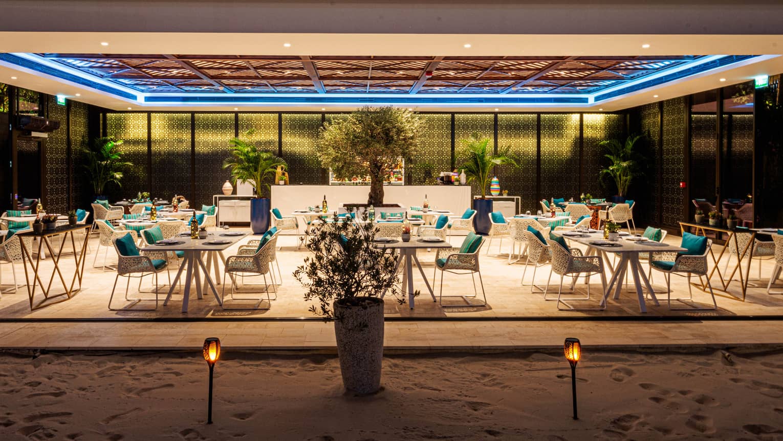 Nighttime view of luxury hotel restaurant dining room open to the beach on one side and surrounded by glass windows on the other sides with a tray ceiling above featuring an inset blue light