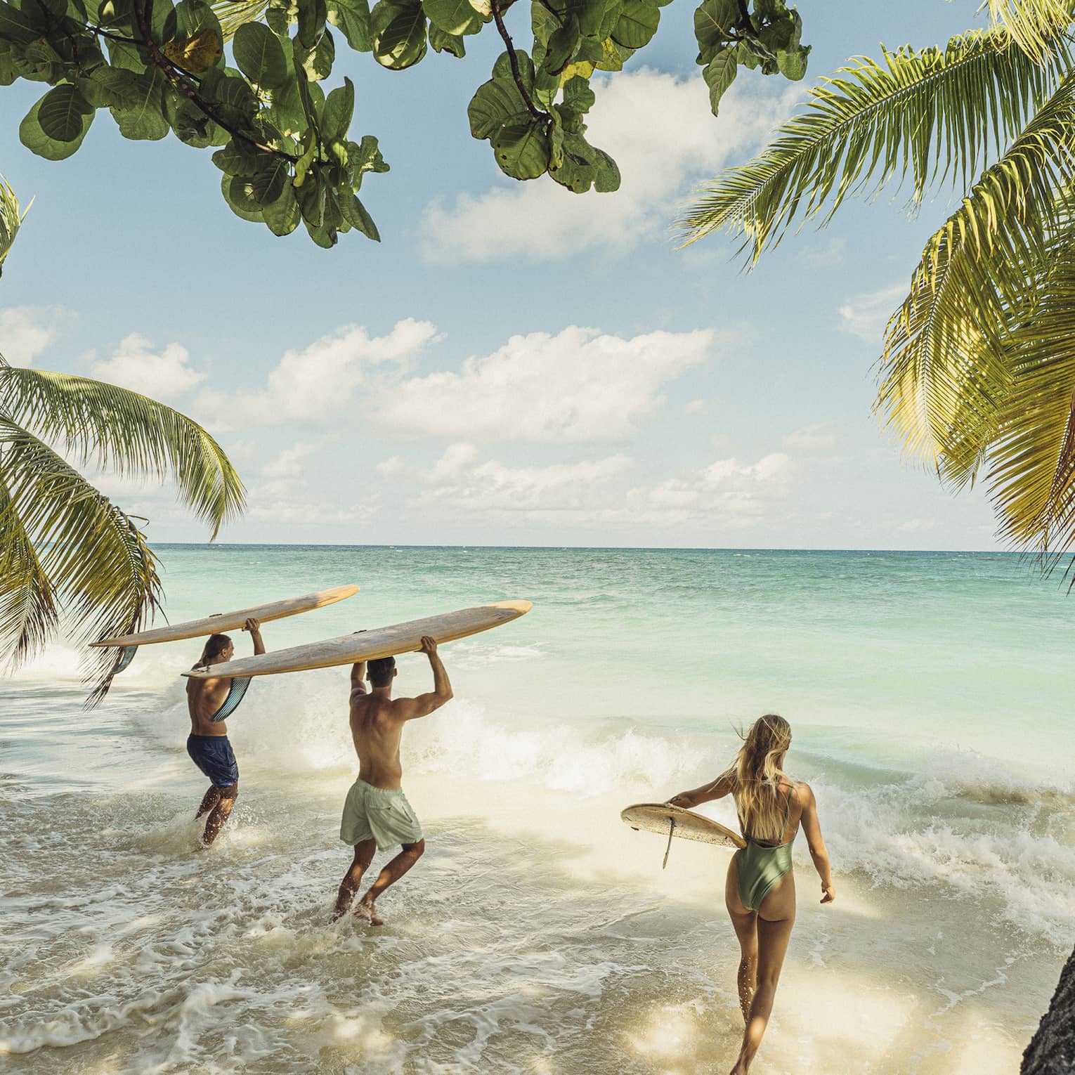 Palm trees extend over waves rushing a sandy beach, as a group of surfers with their boards bound toward the turquoise sea.
