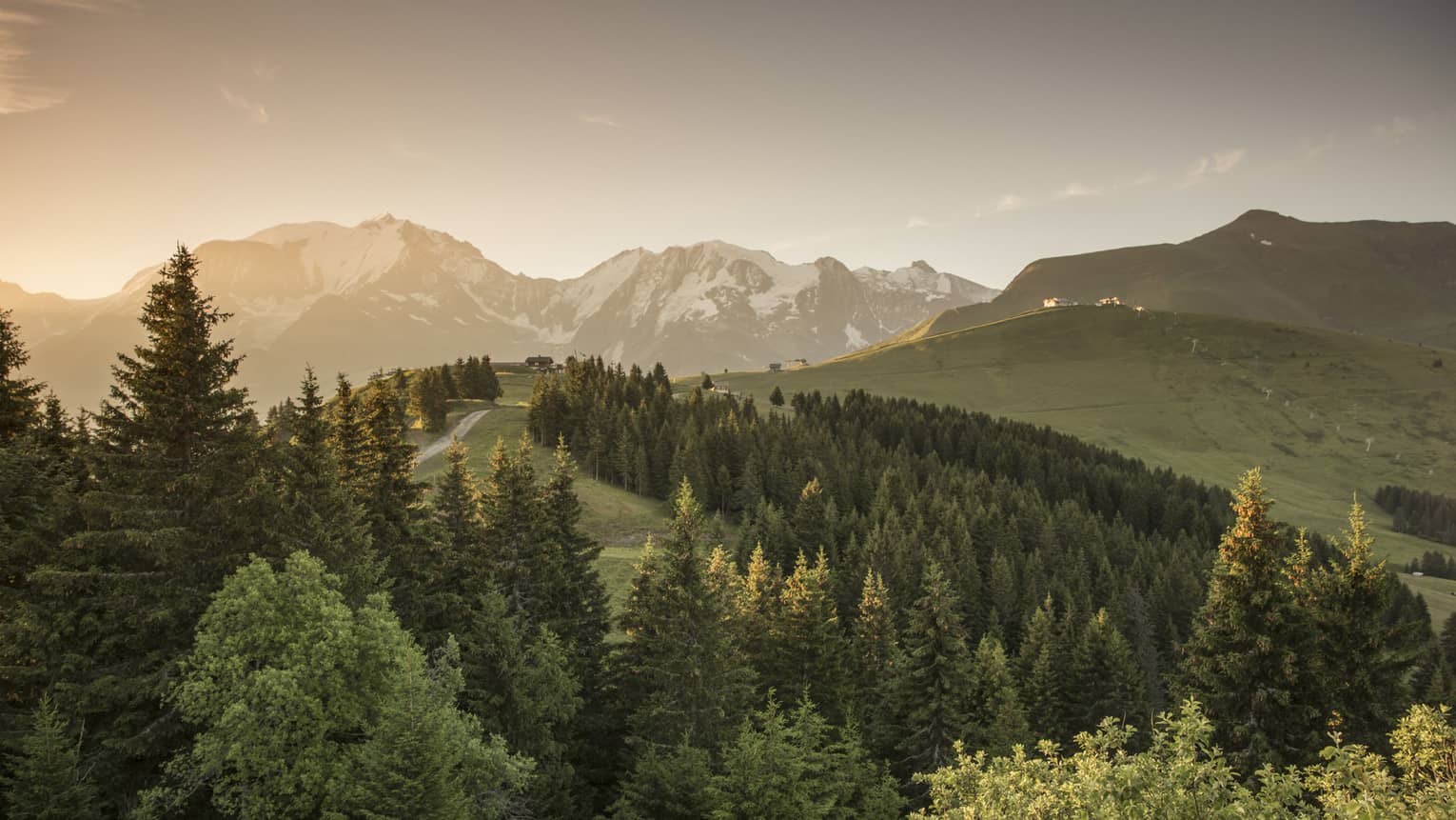 Sun sets over snow-capped mountains, green forests, trails in French Alps