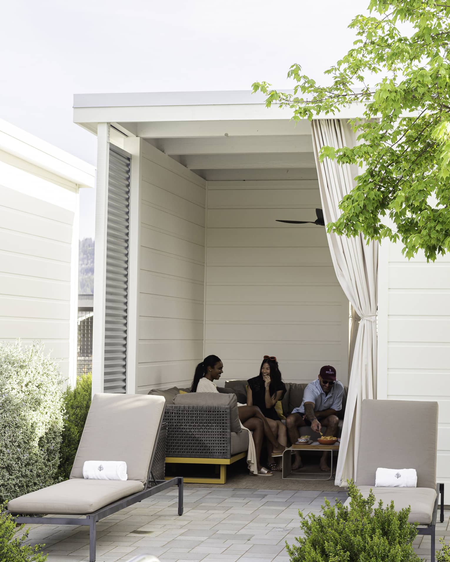 Three guests eat snacks on a large couch in a square cabana with a tied-off curtain and two lounge chairs at the entrance.