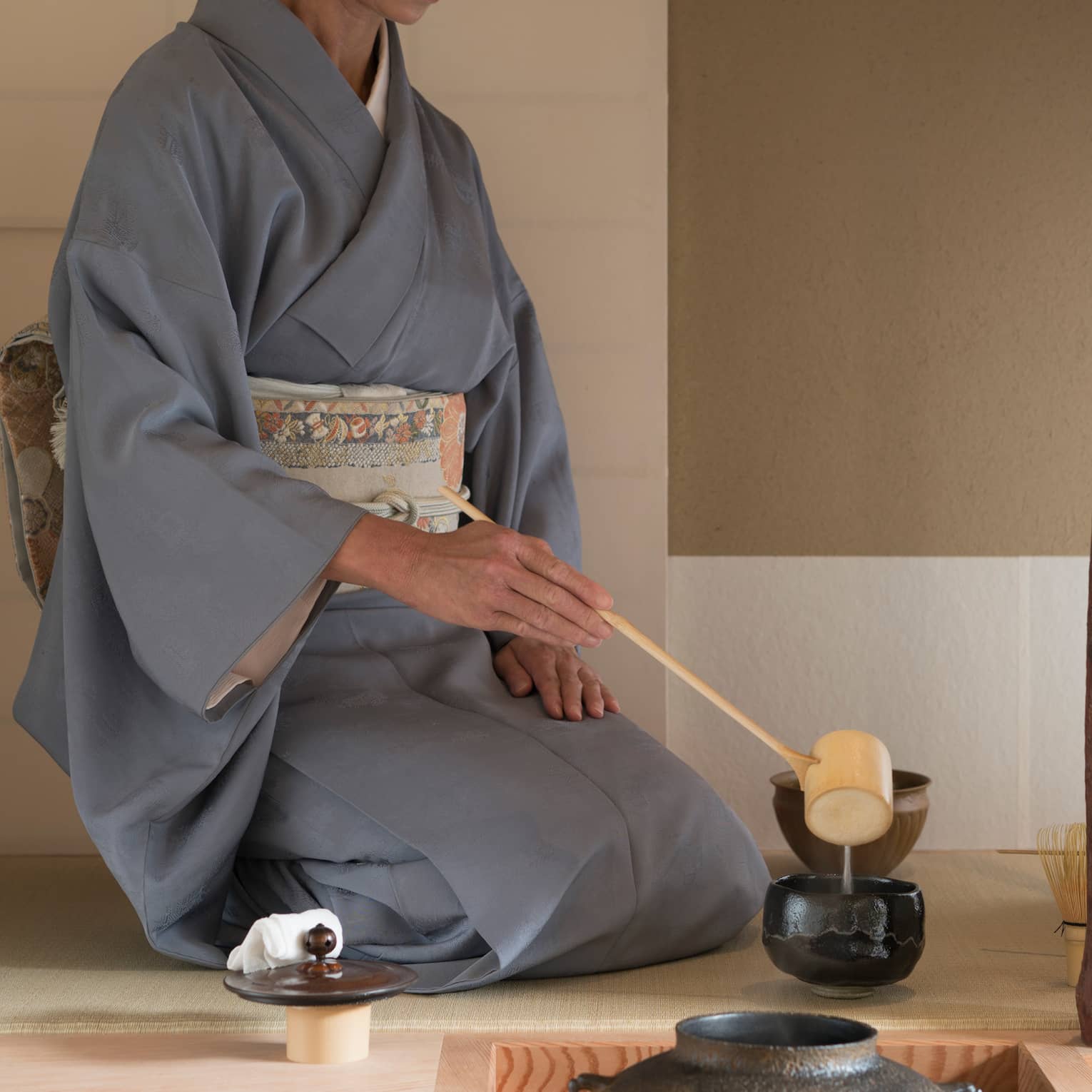 Woman in robe kneels on floor, ladles tea into cup with wood spoon