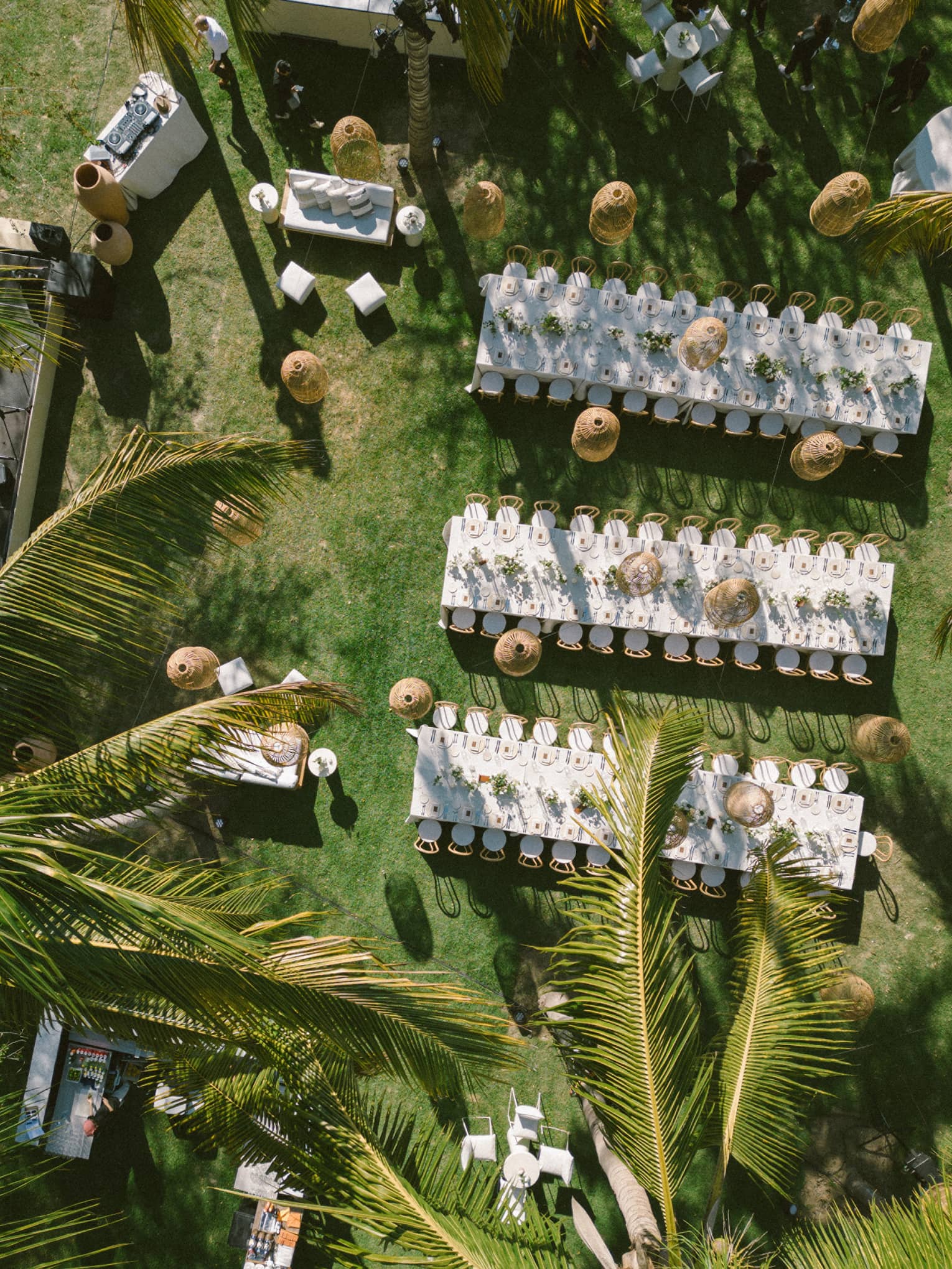 An aerial view of an outdoor event setup on a green lawn surrounded by palm trees. Setup includes long dining tables set for an event, chairs, lighting and other decor.