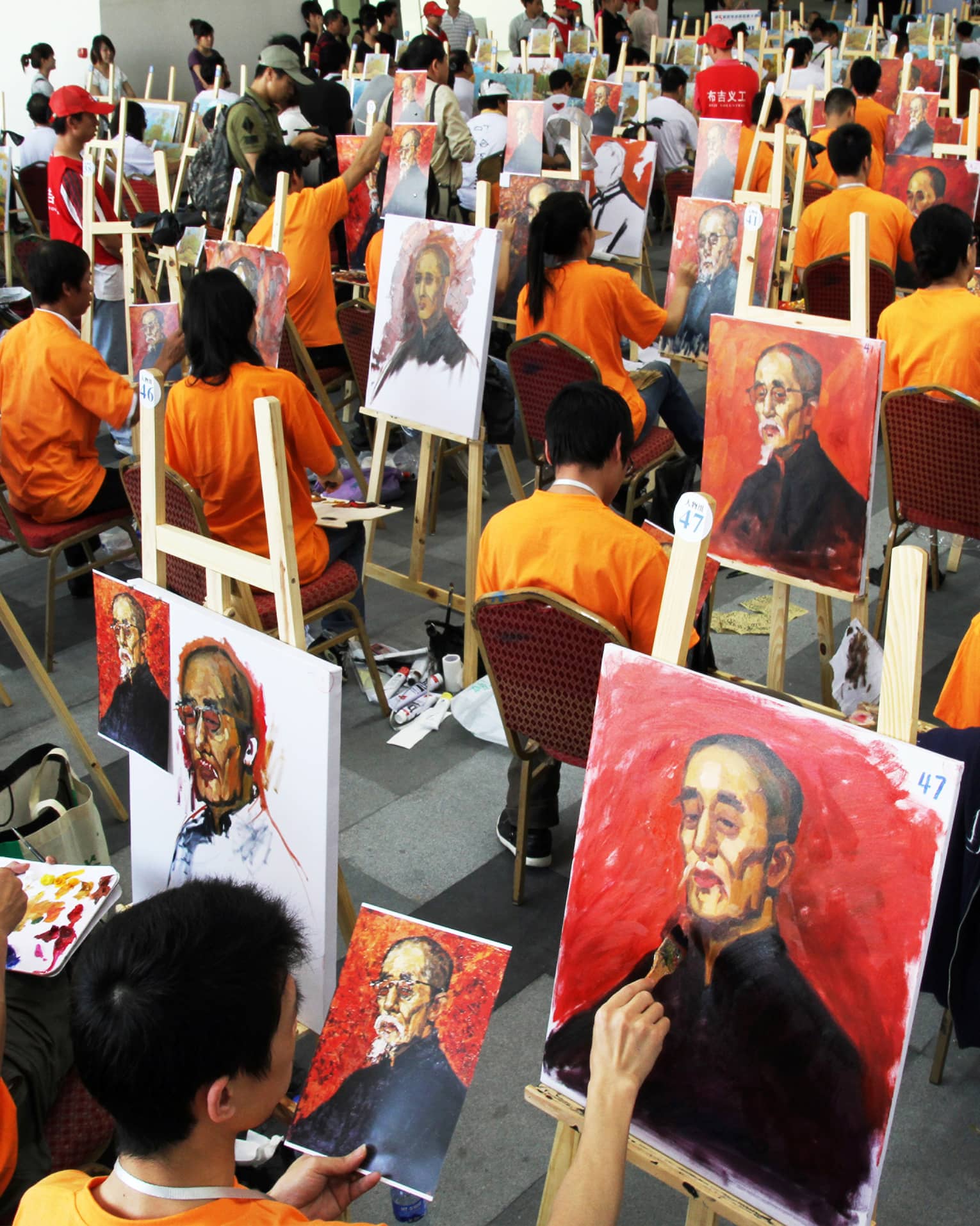 Dozens of people wearing orange shirts sitting at easels and painting the same portrait.