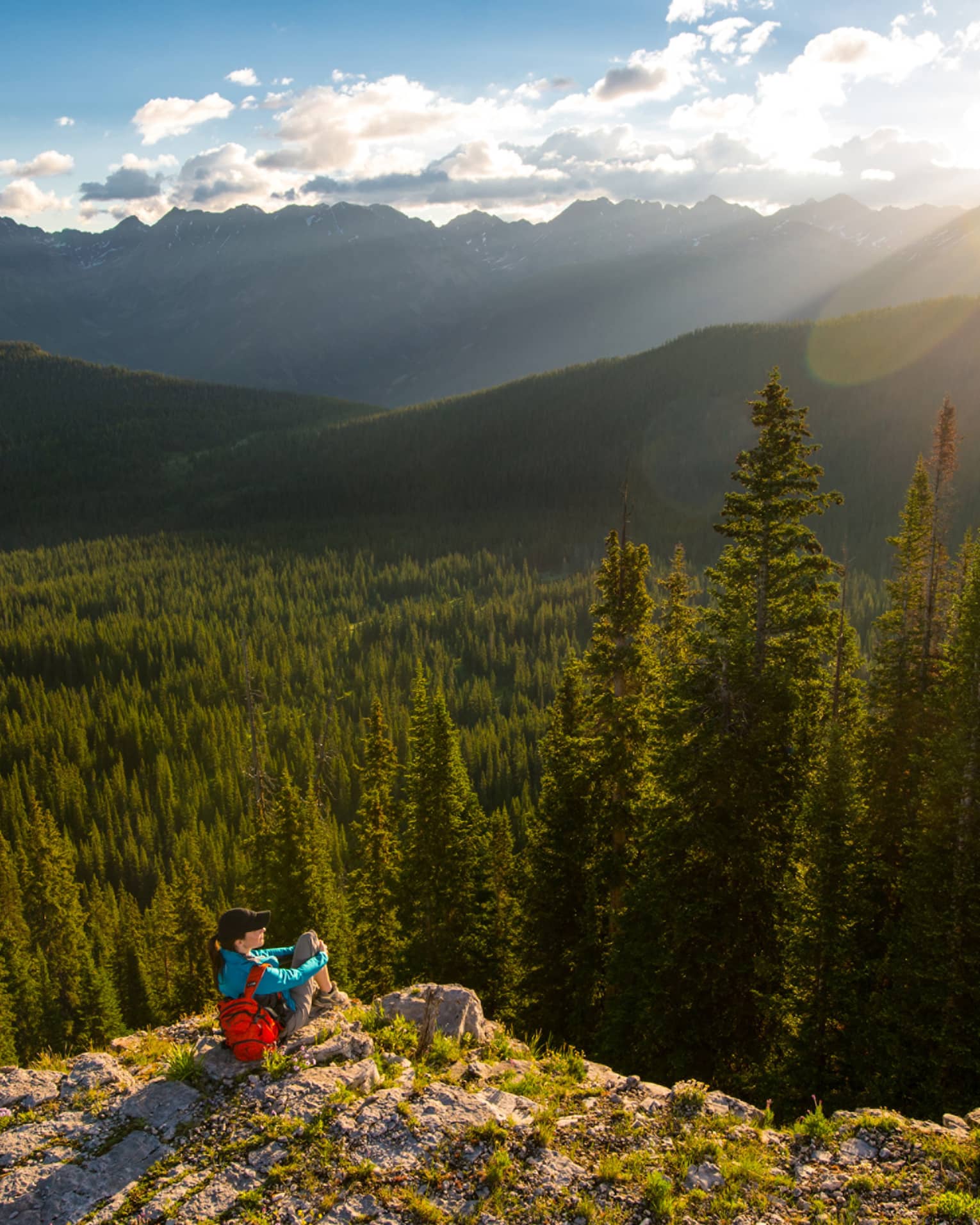 Person sits at edge of cliff overlooking trees, mountains as sun starts to set
