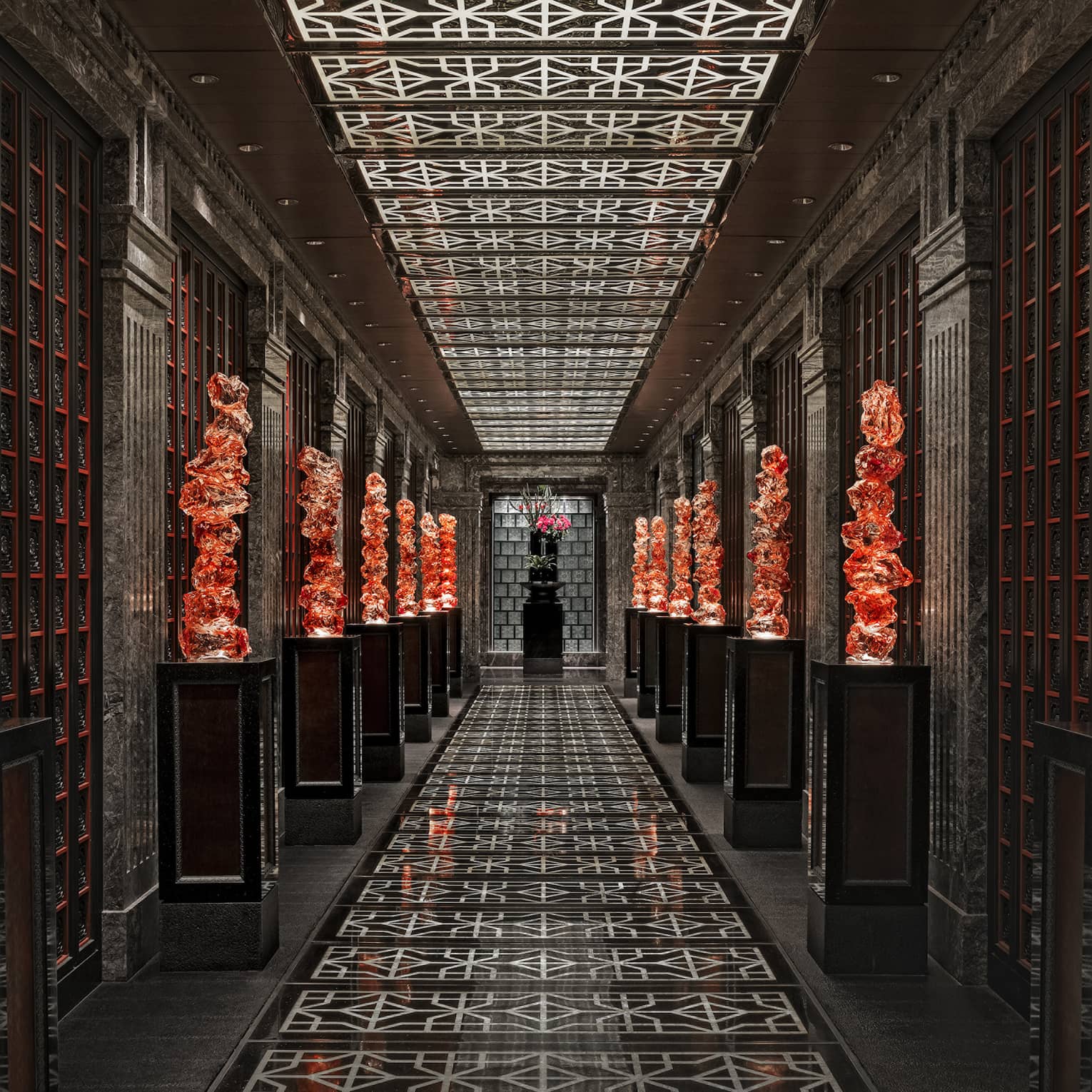 View down a long black-tiled hall lined with orange glass sculptures on pedestals, the geometric ceiling mirroring the floor.