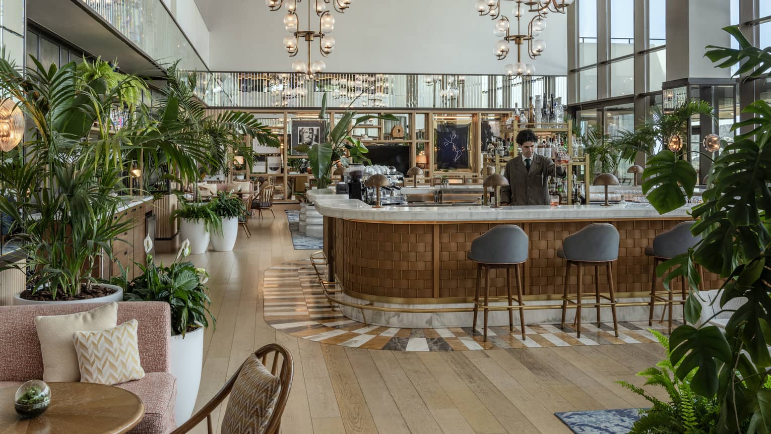 A person stands behind the bar in a spacious lounge with a wood floor, modern lighting, large potted plants, seating and tables, and a wall of windows admitting natural light