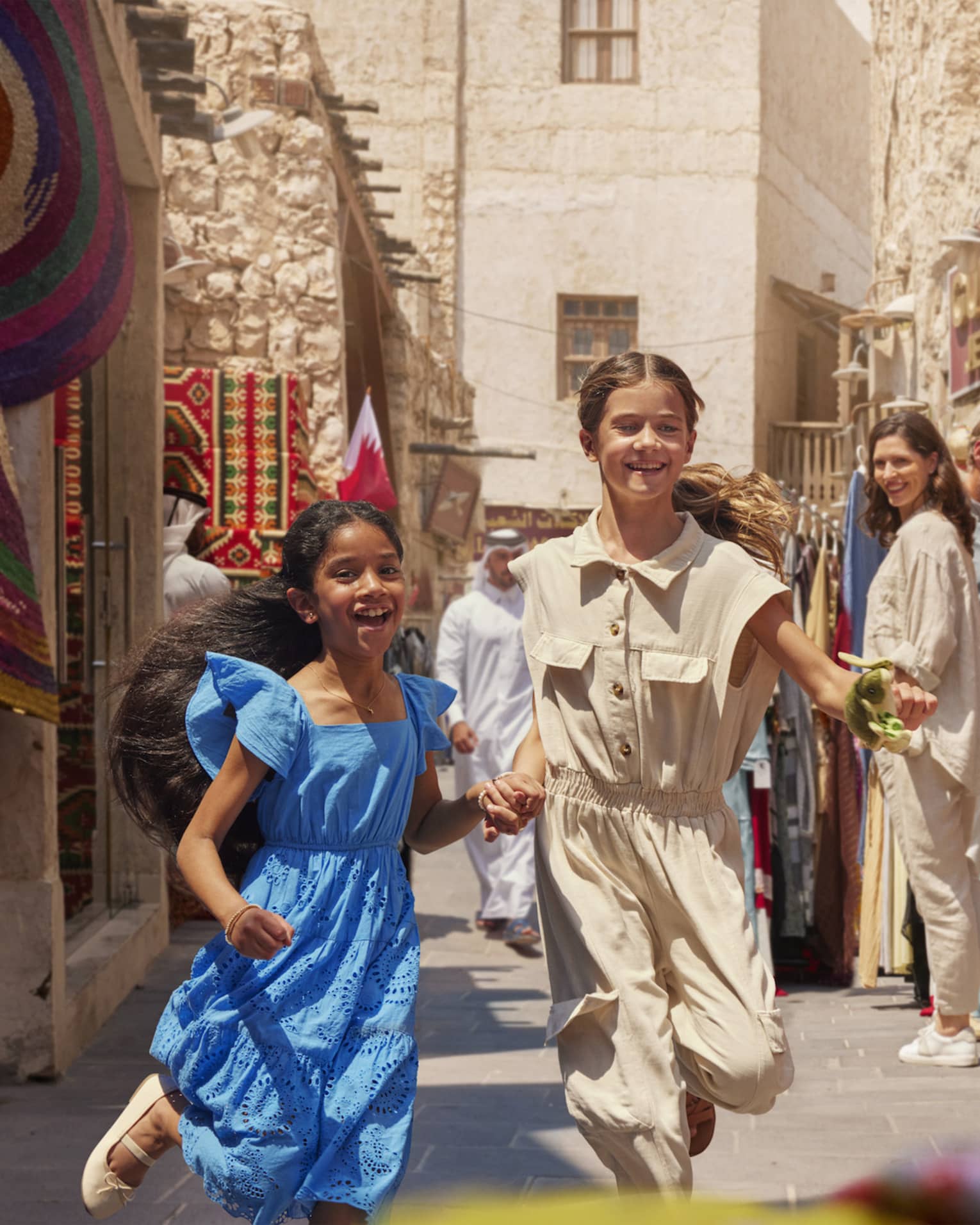A busy, sunny open-air market between sandstone buidlings; two children run among shoppers browsing vibrant clothing.