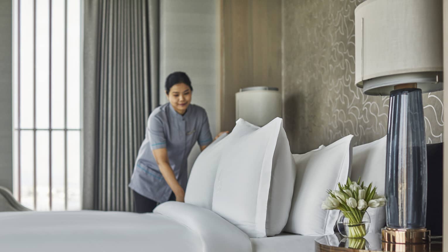 A room service attendant fluffing pillows on a freshly made bed