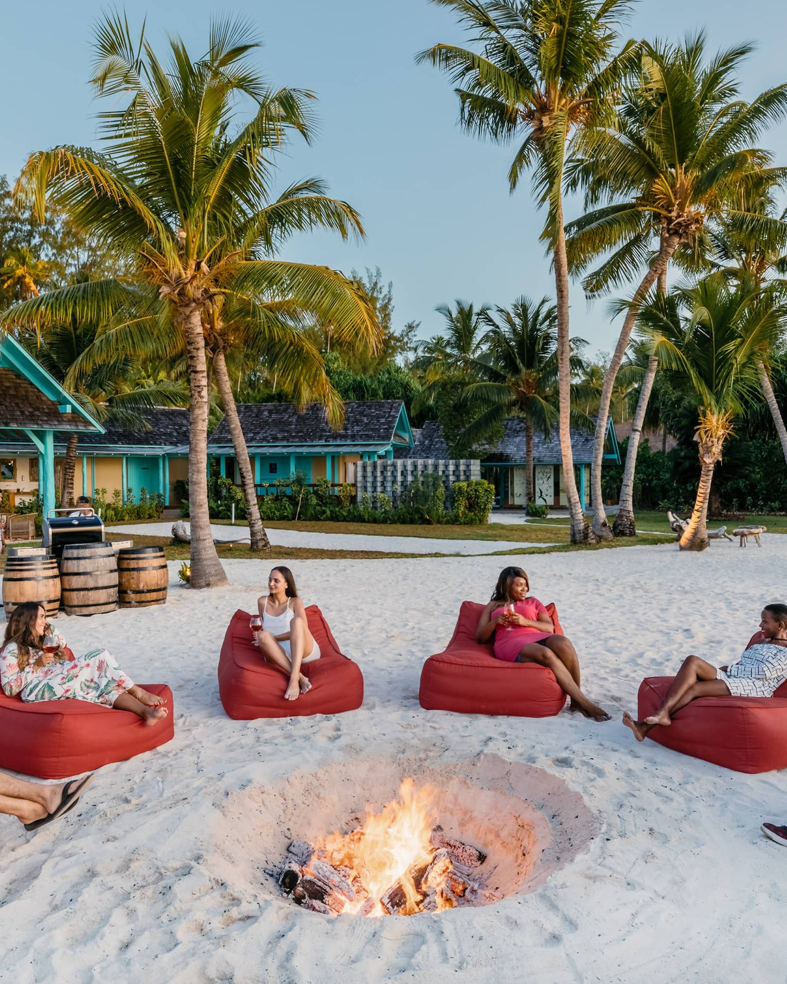 People seated in semi-circle of red bean bags around firepit in sand with palm trees in backdrop