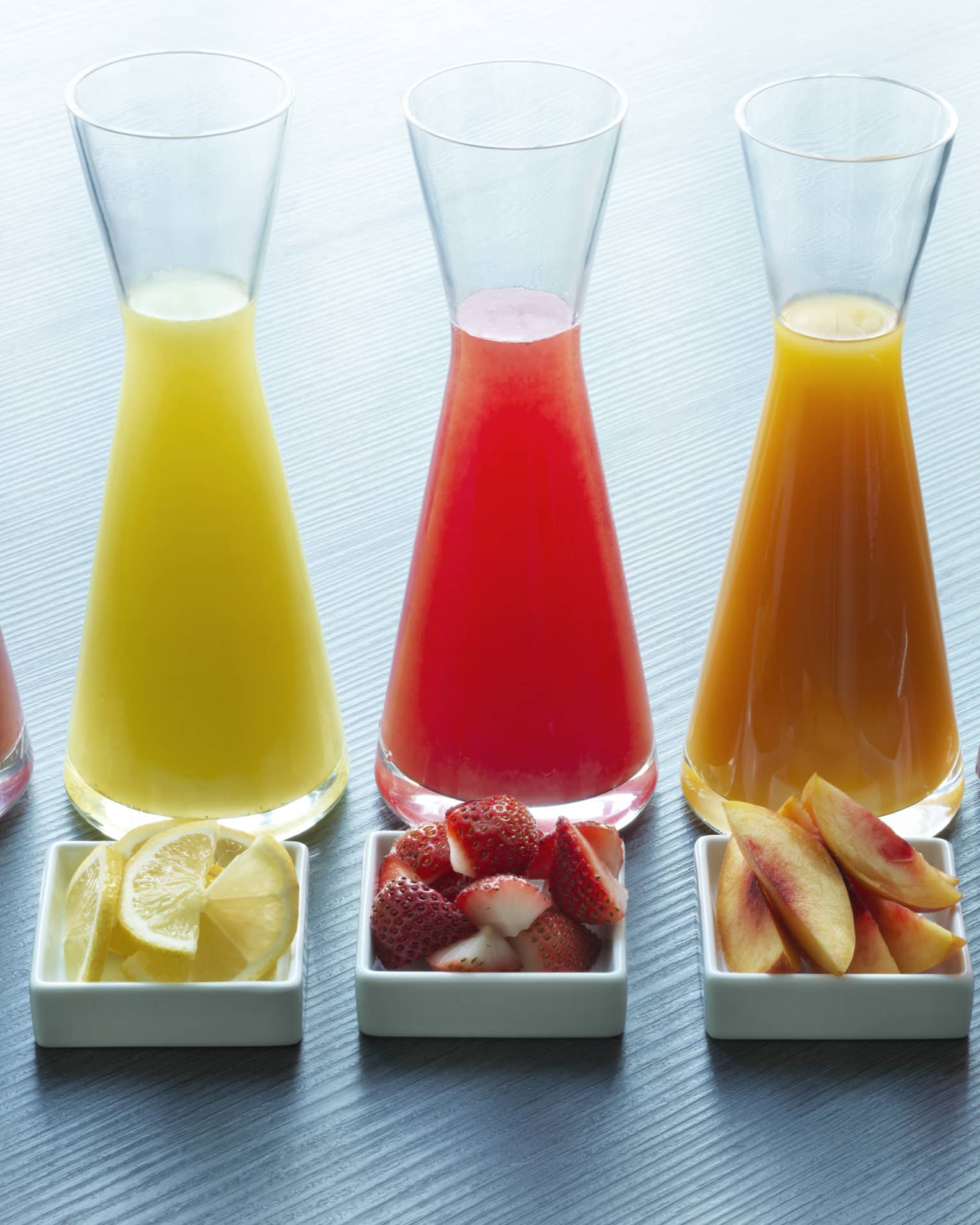 Five glass juice jugs aligned with five small trays of fruit