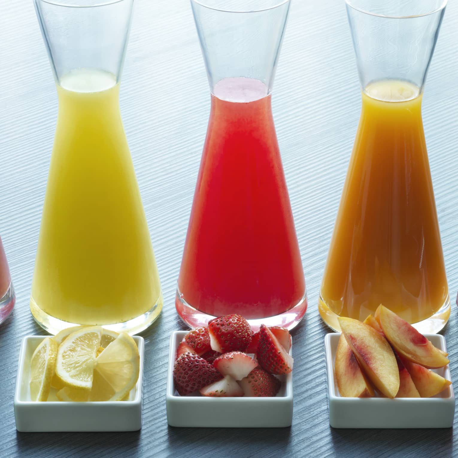 Five glass juice jugs aligned with five small trays of fruit
