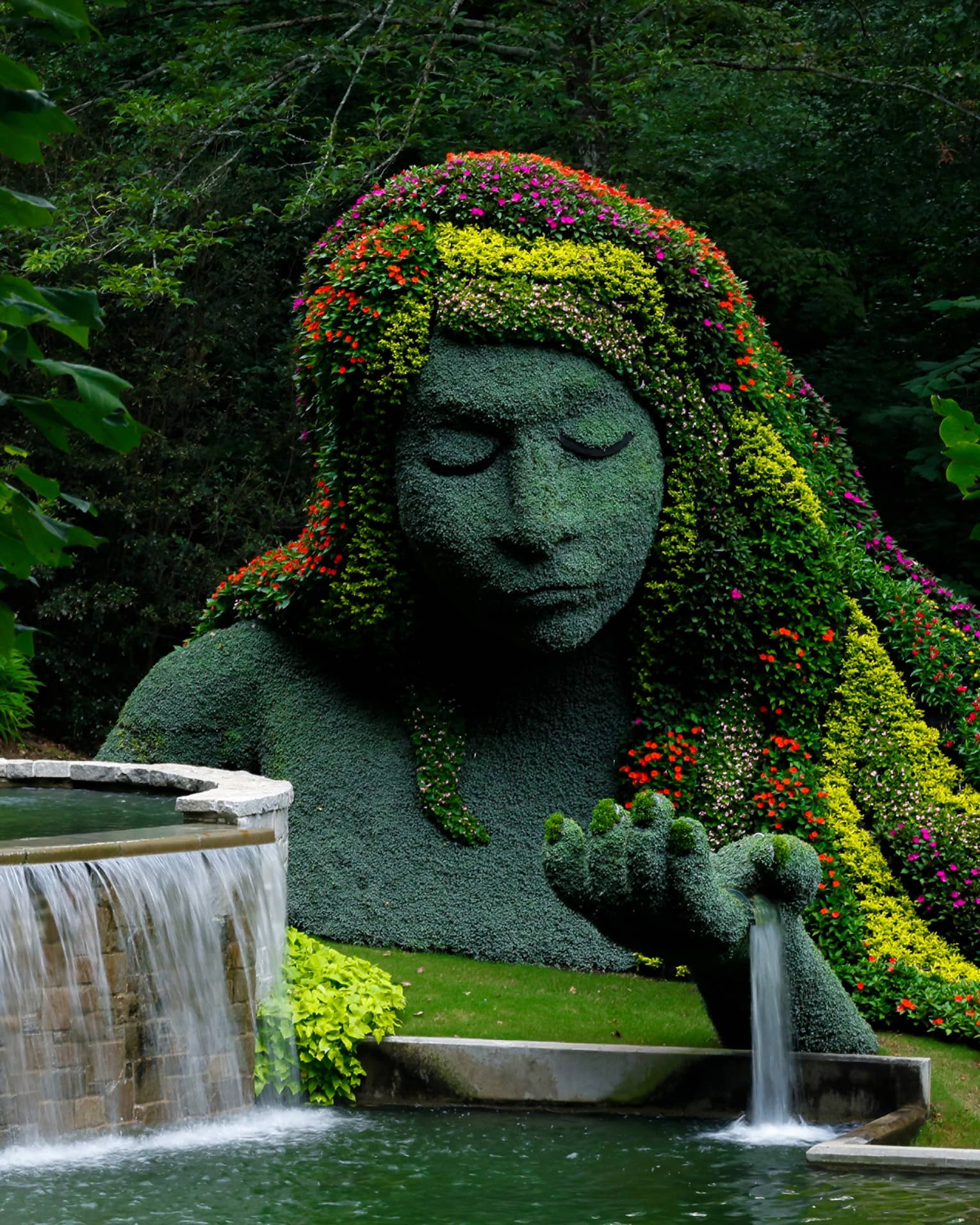 A massive statue covered in pale greenery, its hair a cascade of vibrant blooms; a fountain flows from its outstretched hand.