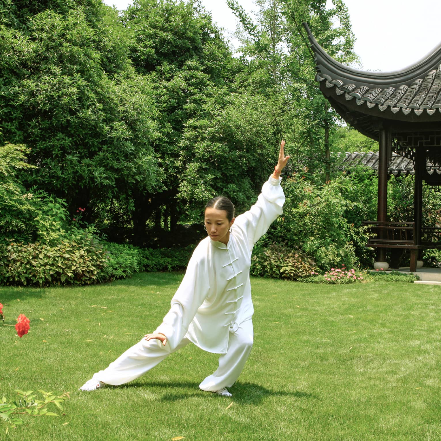 An instructor in a white tai chi outfit, arms and legs outstretched in a classic pose, in front of a pagoda-shaped gazebo.