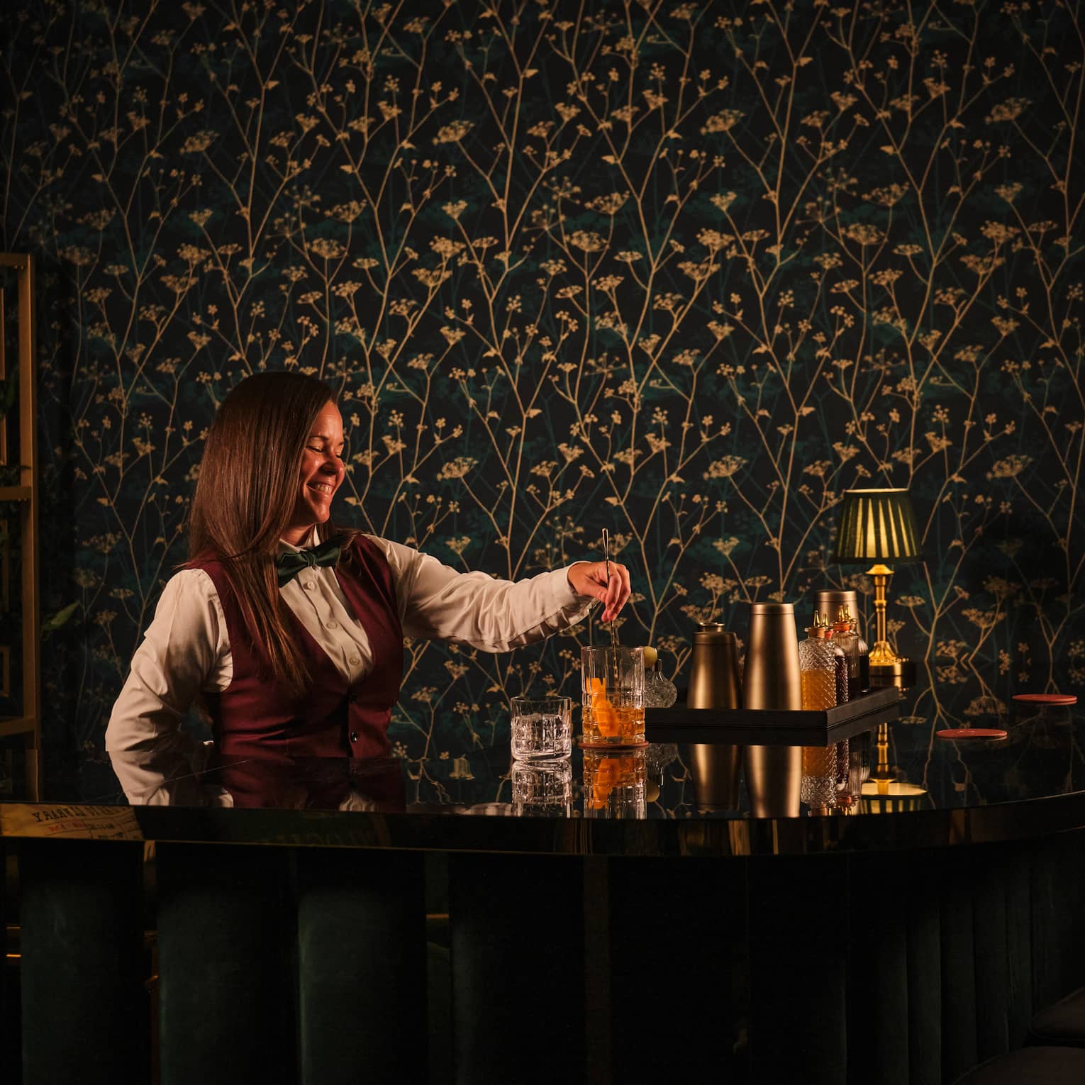 Bartender wearing white shirt, burgundy vest and black bowtie pours a cocktail from behind the bar