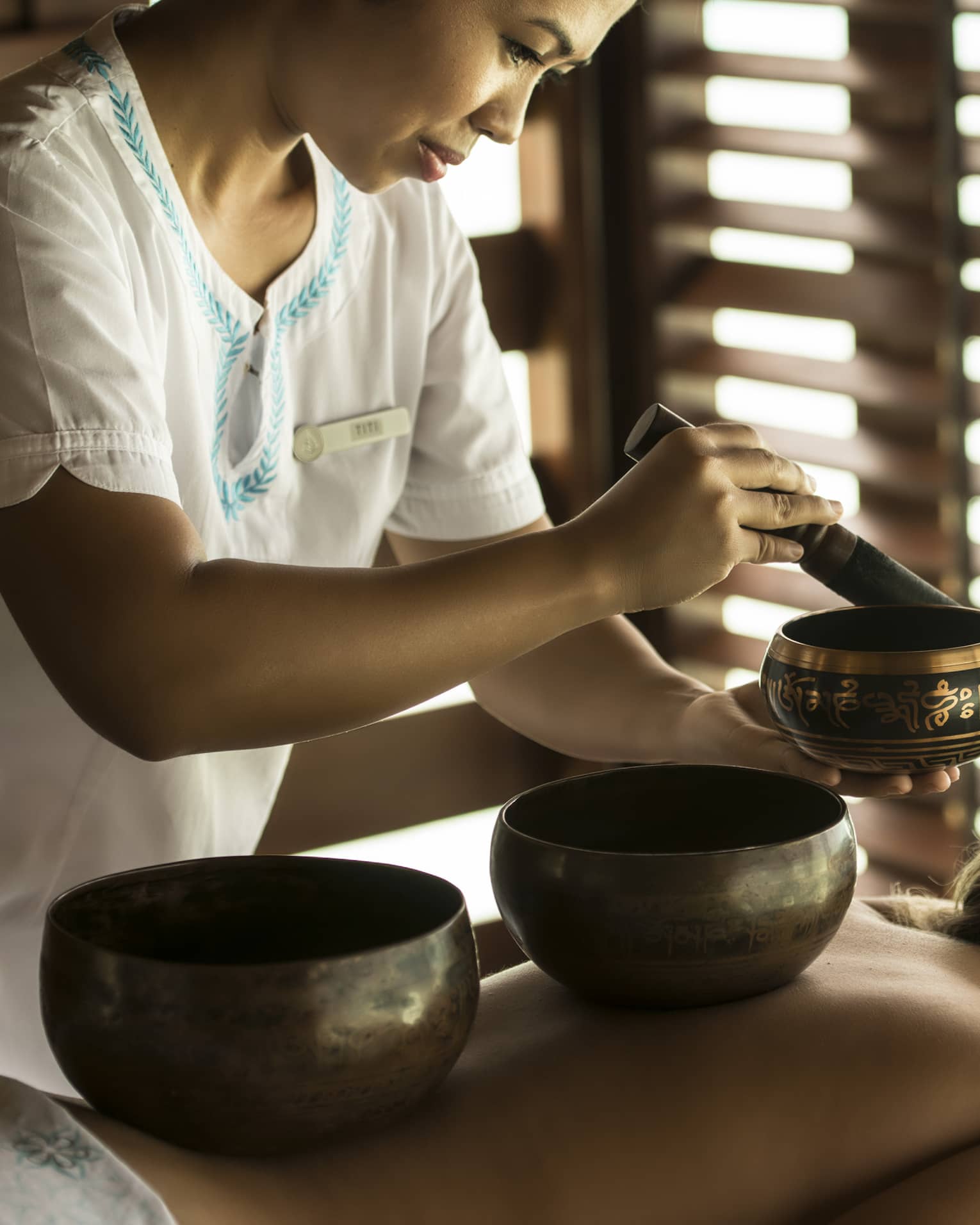 Guest with singing bowls on bare back lies on treatment table which sound specialist plays another singing bowl during a sound healing session