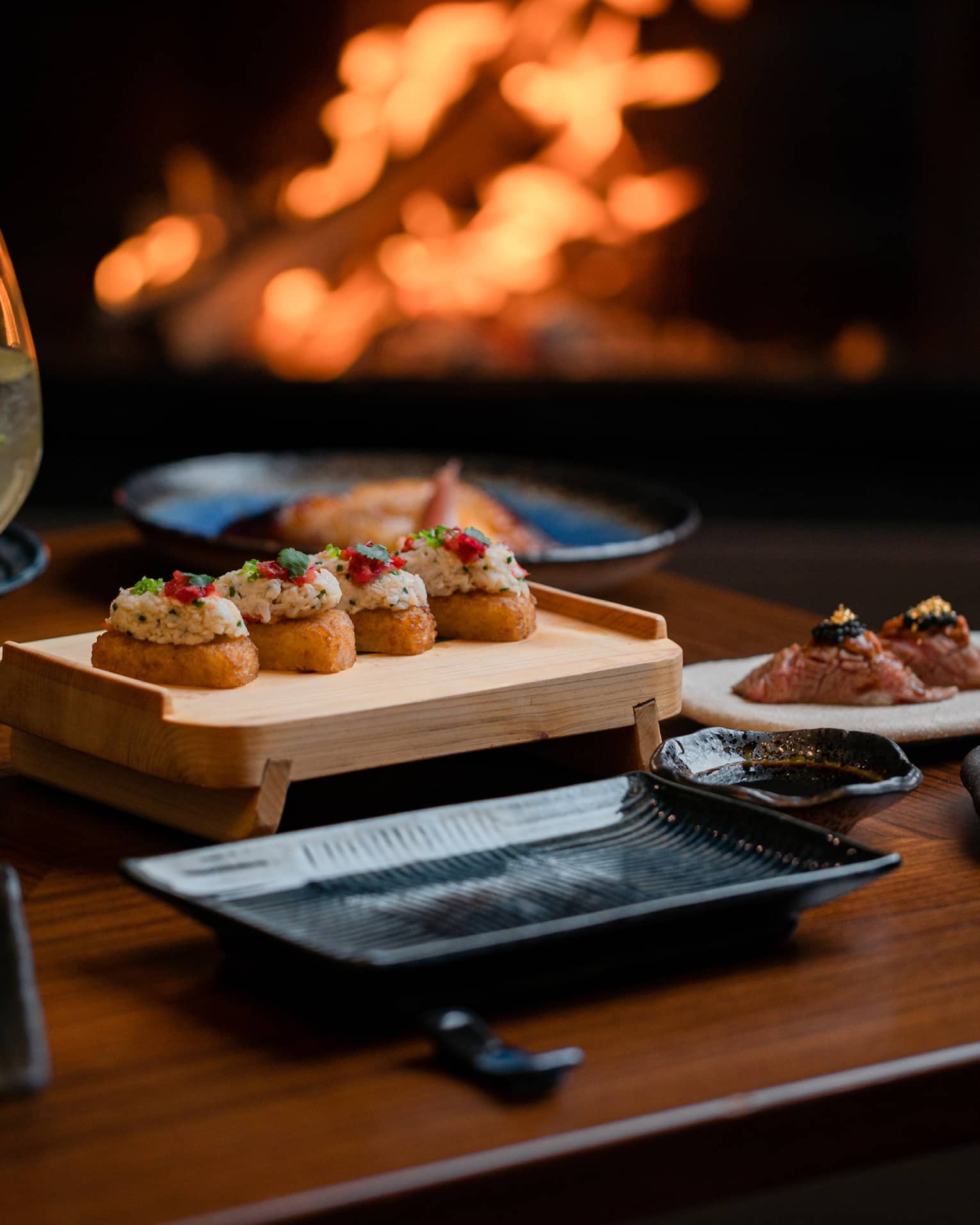 A cocktail and appetisers placed on a wooden table with a fireplace glowing in the background, creating a cosy dining atmosphere