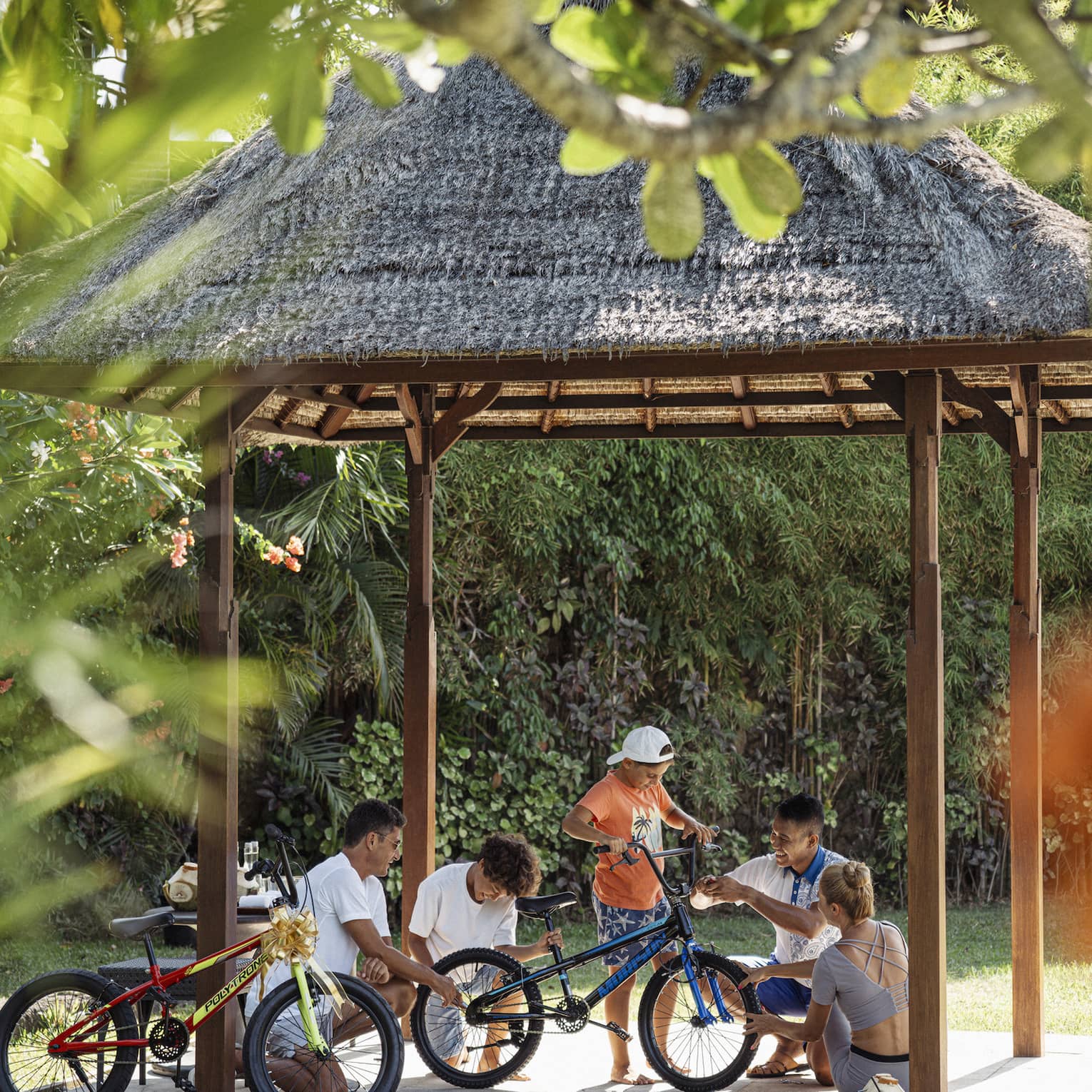 View through foliage of four adults and a child working on a bicycle under a thatched hut surrounded by lush greenery.