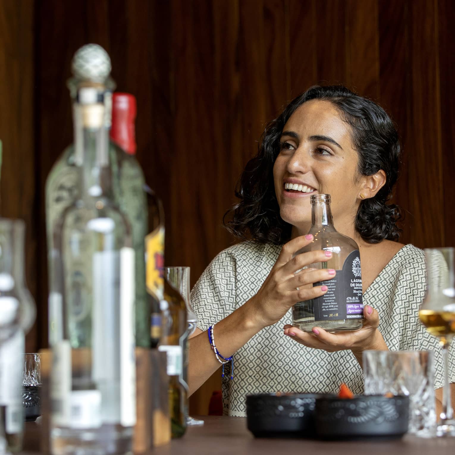 A guest cradles a short, wide bottle of liquor at a table littered with bottles of various sizes and grappa glasses.