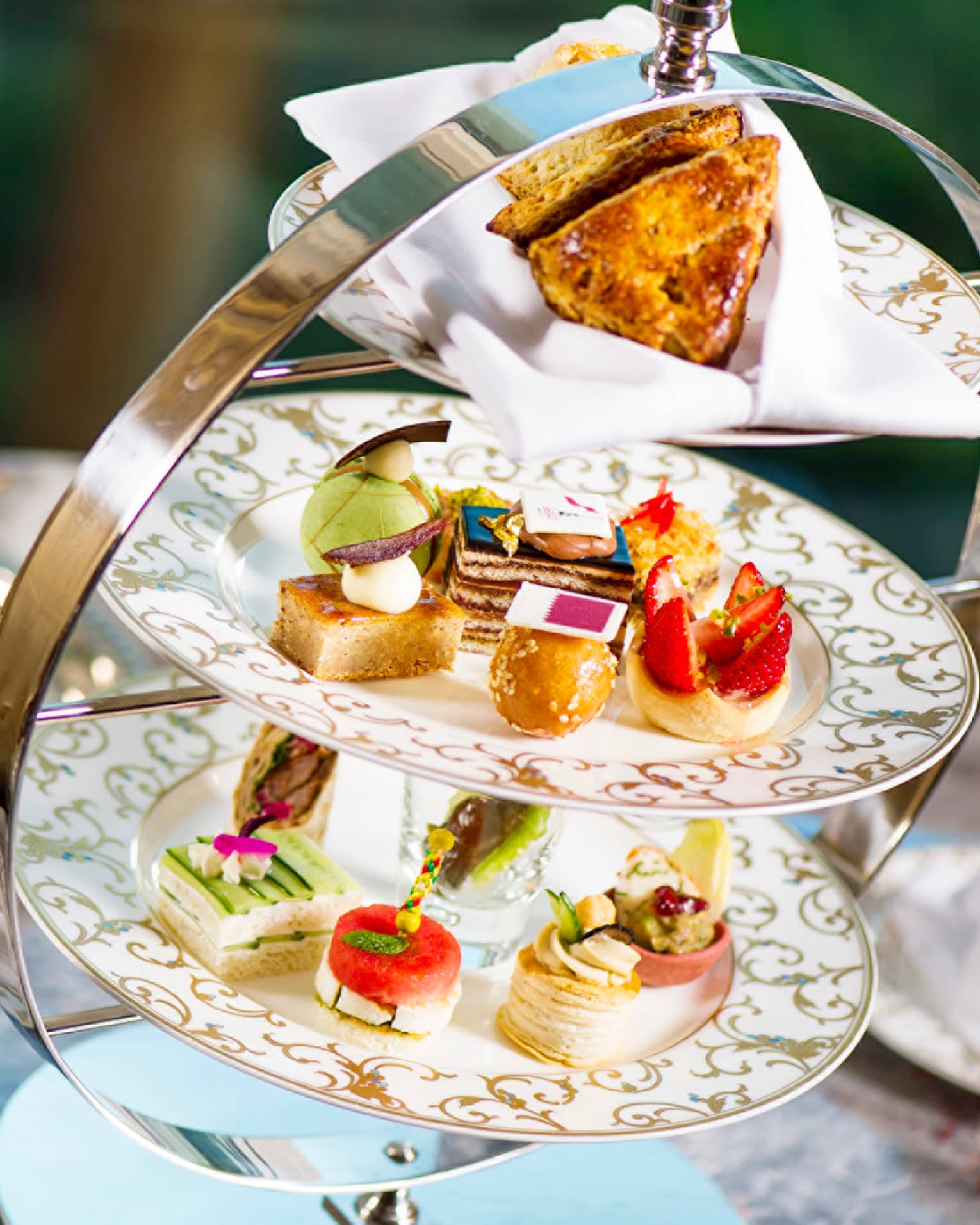 Small, artful pastries displayed on gold-accented china plates in a three-tier stand. Behind, a silver teapot and tray.