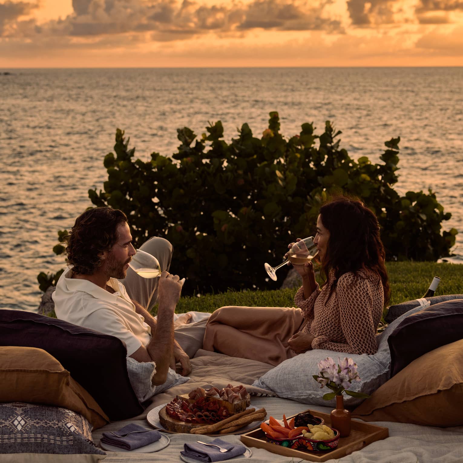 A man and woman lay on blanket and pillows during luxury picnic at sunset, next to the ocean