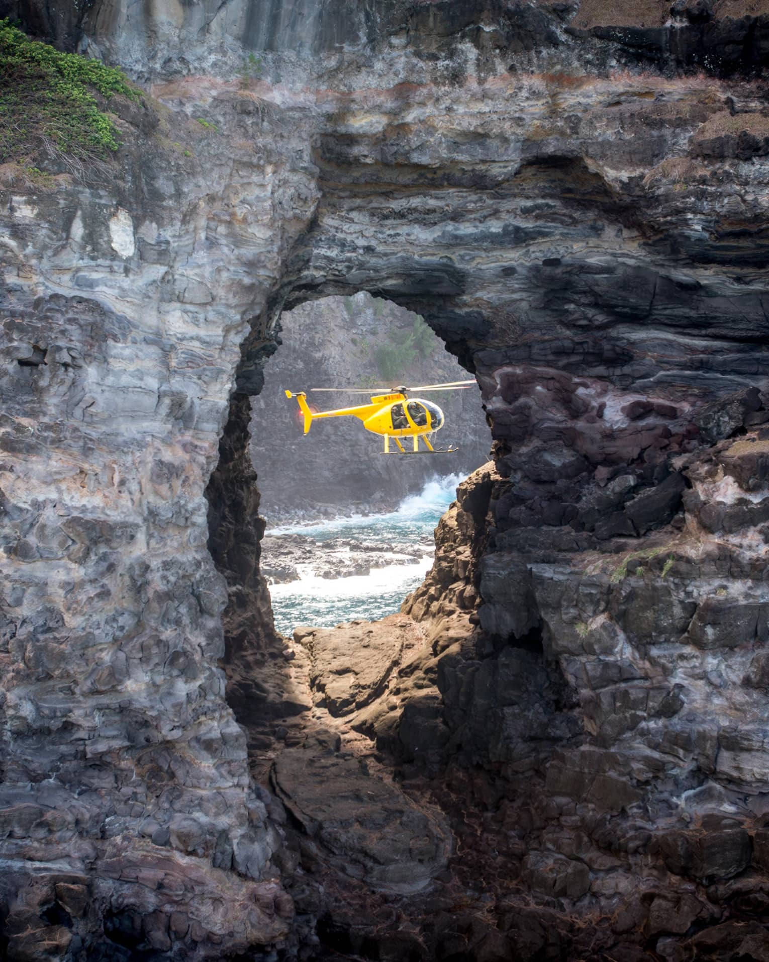 A sideview of a yellow holicopter, seen through an opening in a rock structure. 
