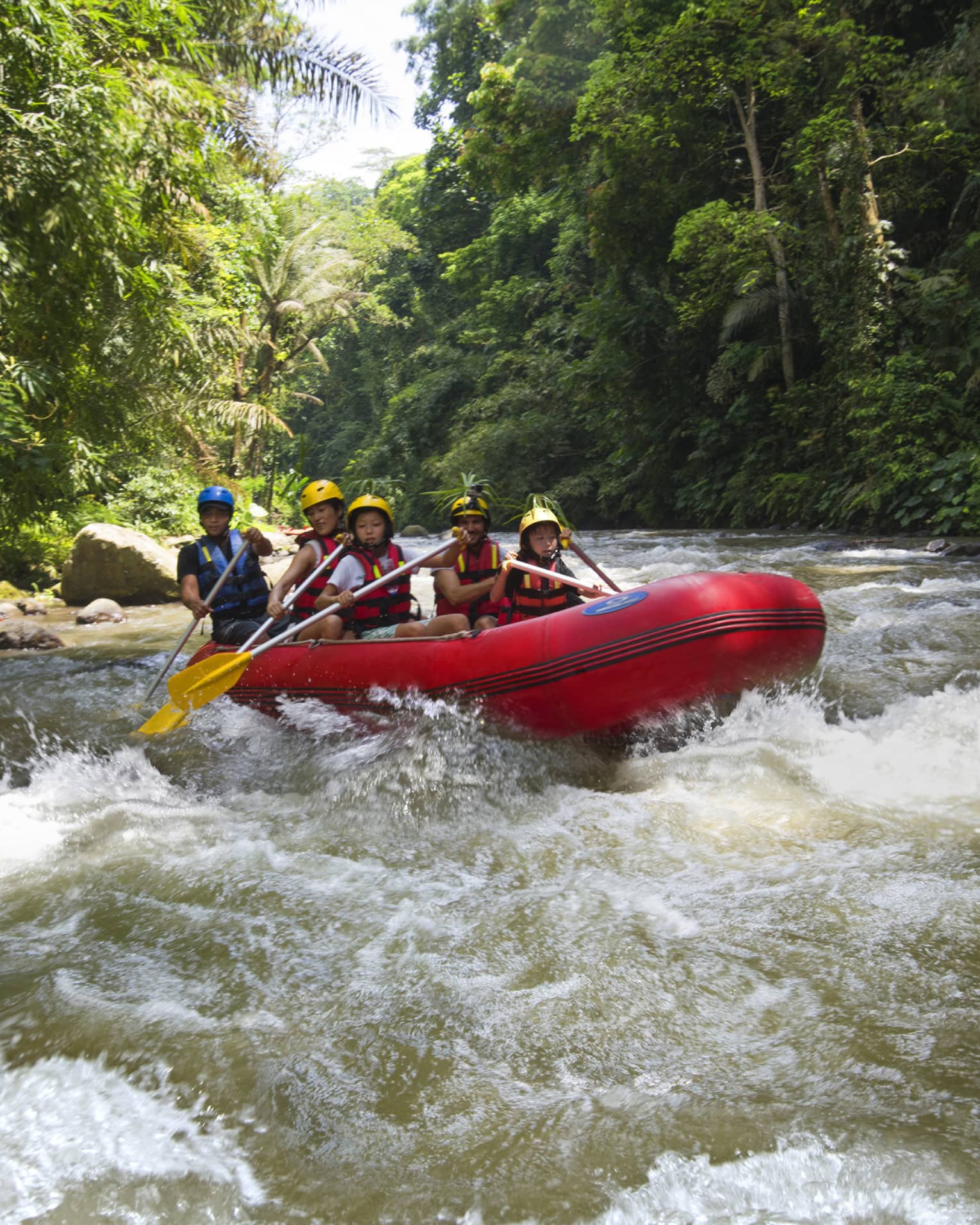 Family of four and guide hold paddles, steer red inflatable raft on rapids in river