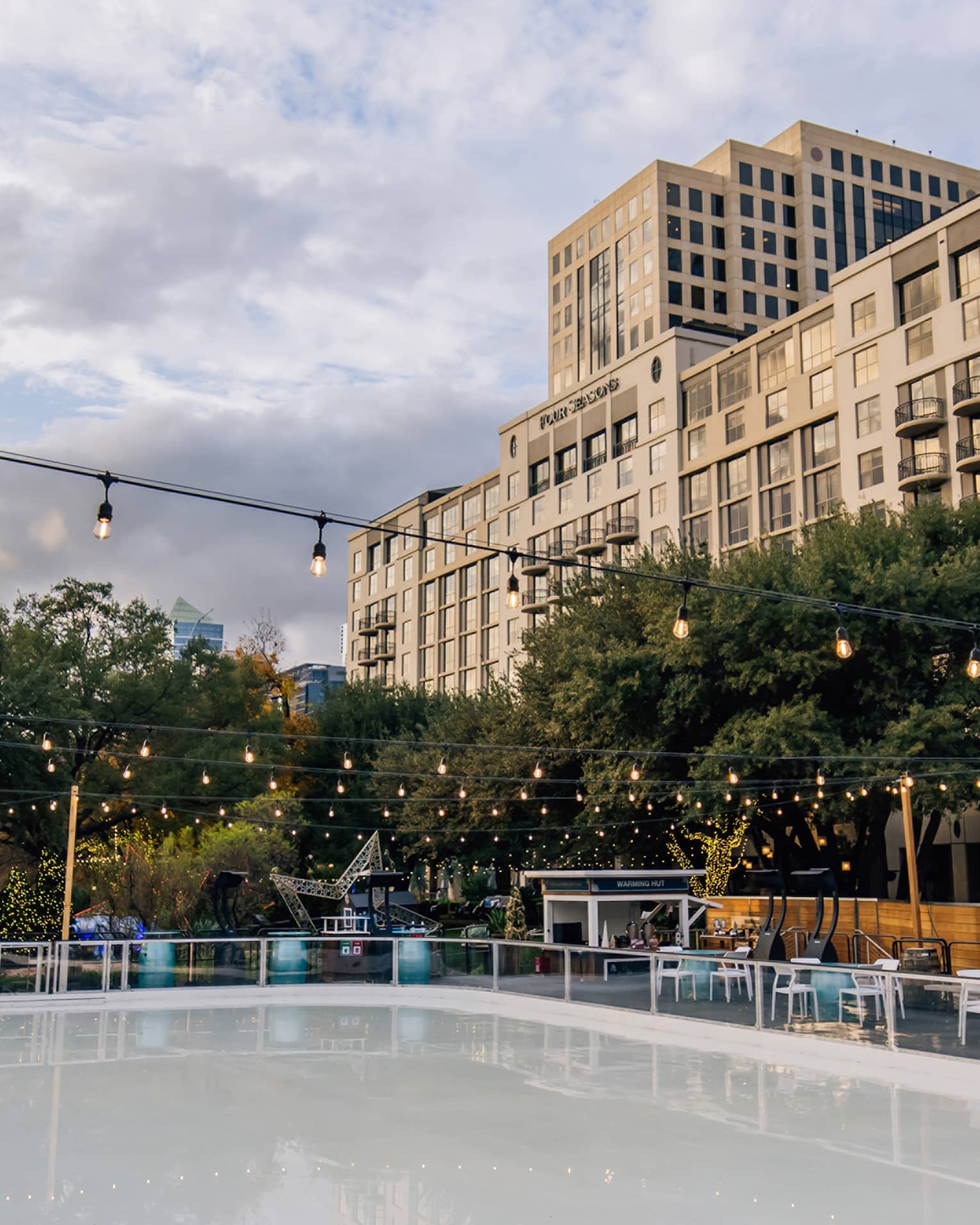 View of the Hotel overlooking an outdoor skating rink surrounded by trees and tables and lit by strings of Edison lights.