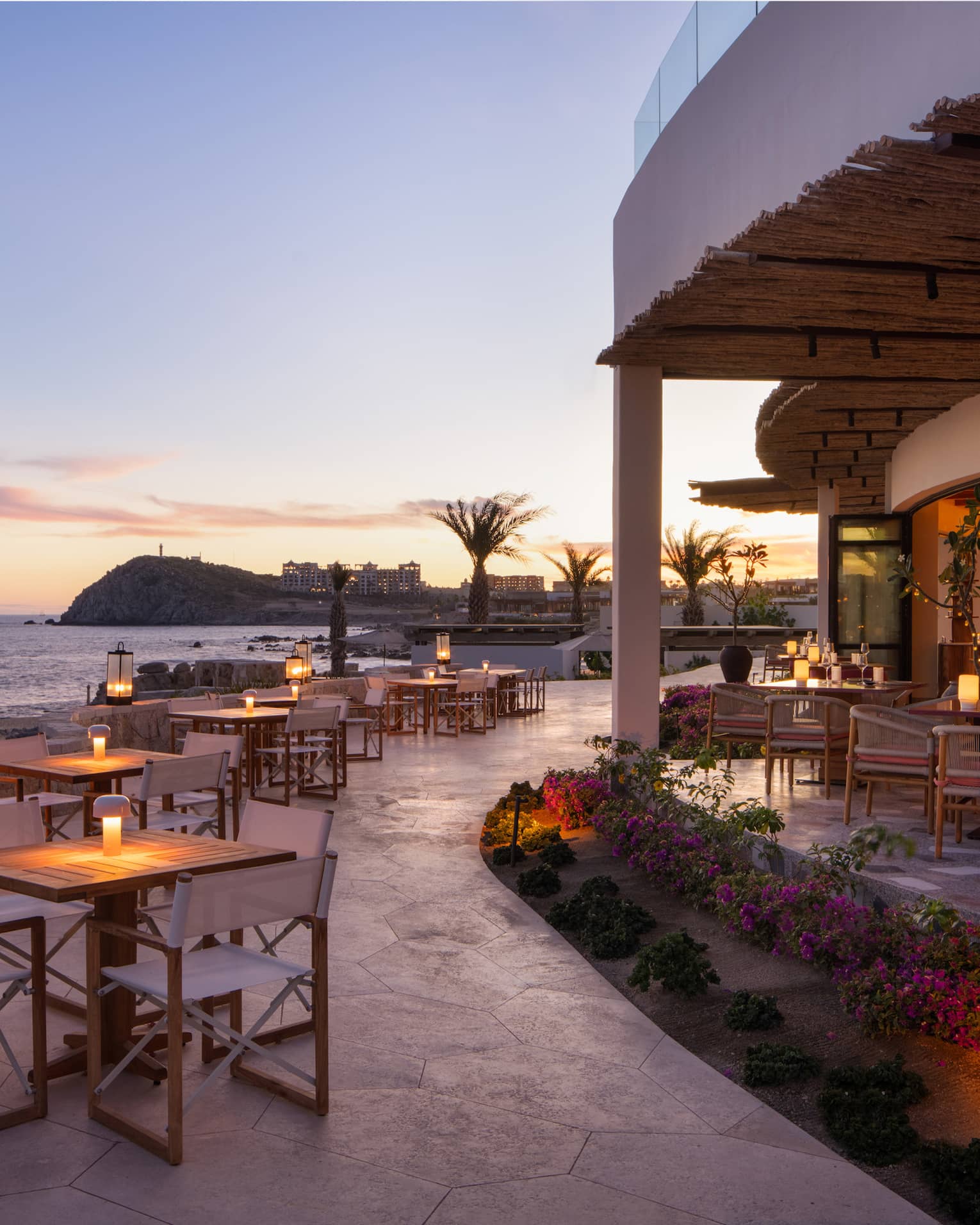 In the warm afterglow of sunset, lanterns light up a large, curved stone restaurant patio at the edge of the ocean.