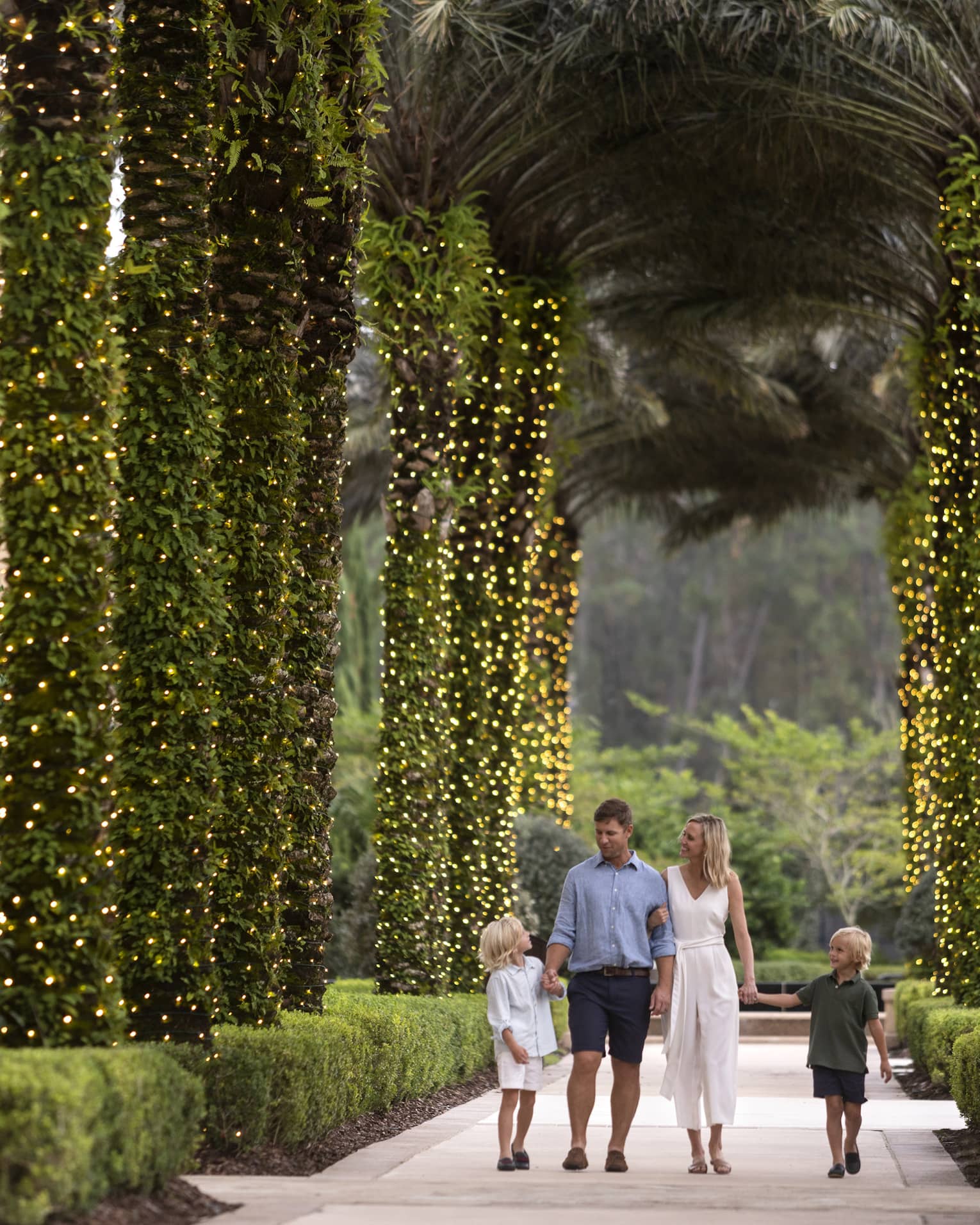 A mother, father and two sons walking along a path of palm trees wrapped in Christmas lights.