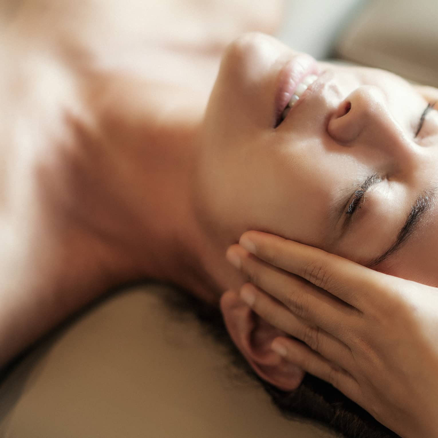 Spa facial, two hands rest on woman's forehead as she closes her eyes, lays under sheet on treatment table