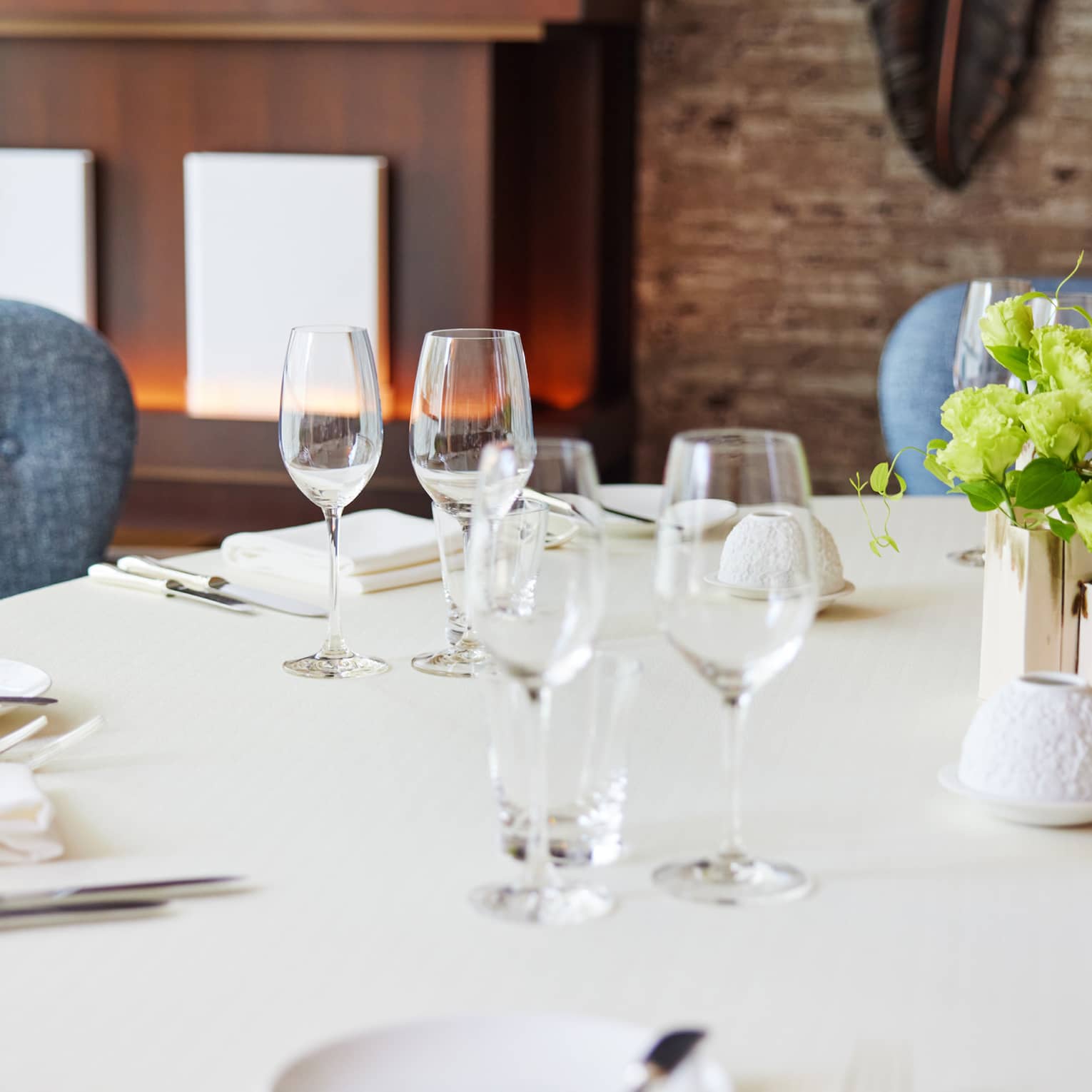 Against a warm backdrop, round table set with white tablecloth, sparkling silver and wine glasses, fresh flowers at centre.