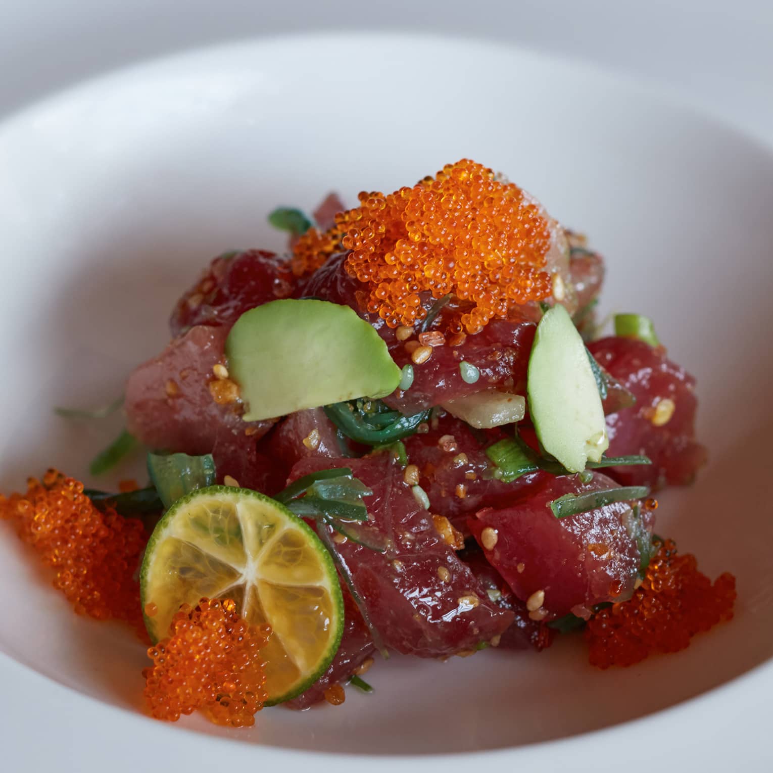 Close-up of tuna sashimi pieces garnished with sesame seeds, avocado, a slice of lime and orange fish roe in a white bowl.