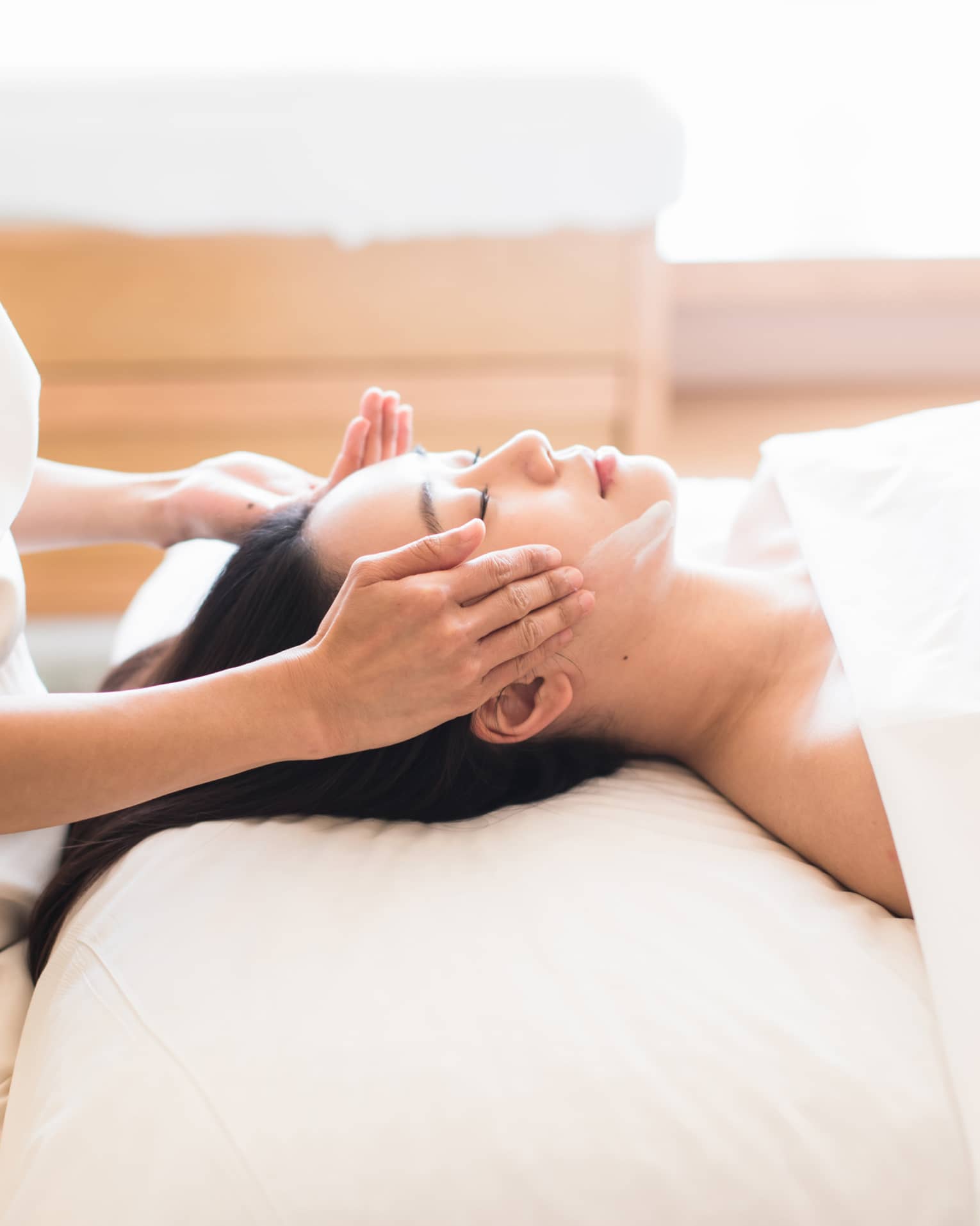 Woman with eyes closed rests head on white pillow as spa attendant rubs cream along her cheekbones