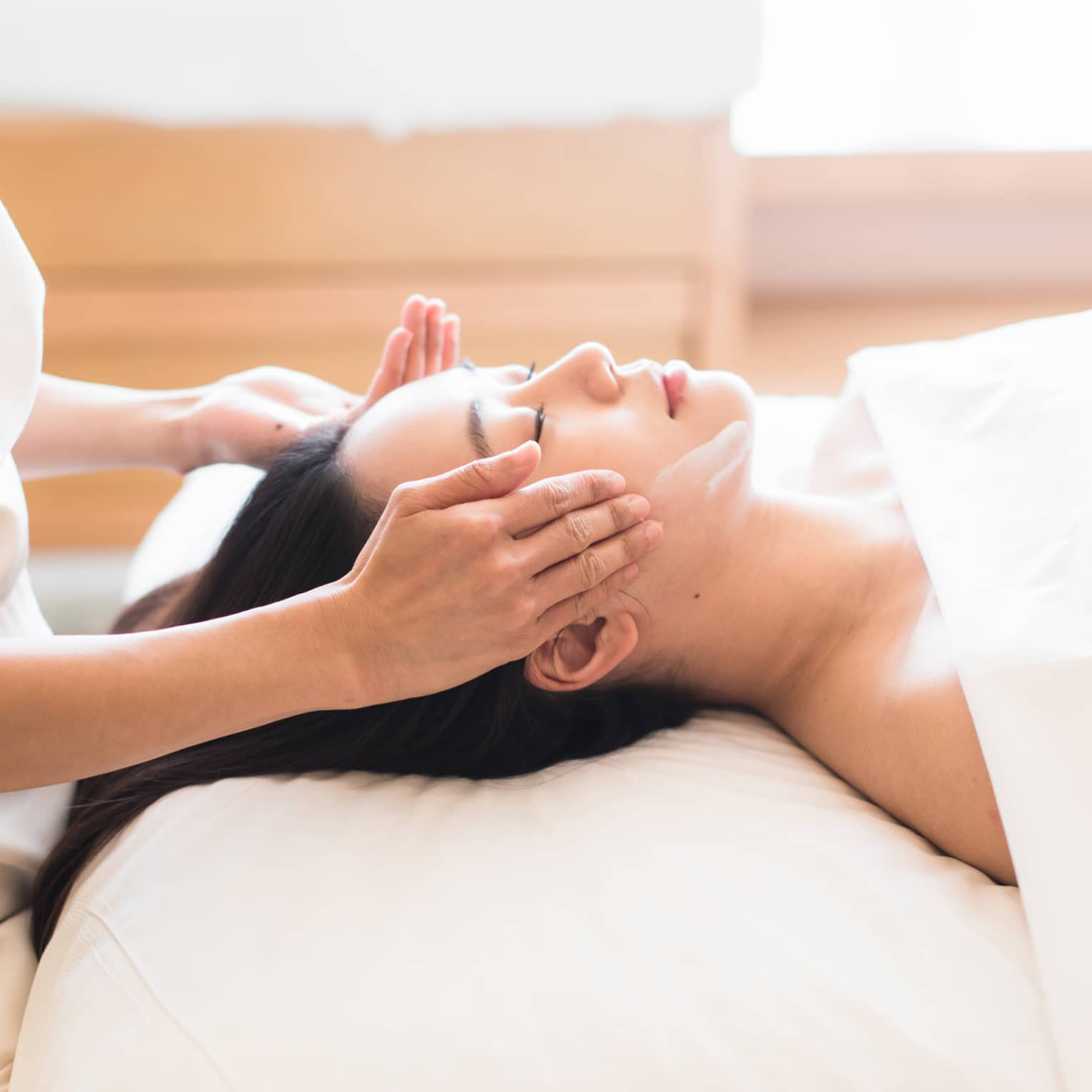 Woman with eyes closed rests head on white pillow as spa attendant rubs cream along her cheekbones