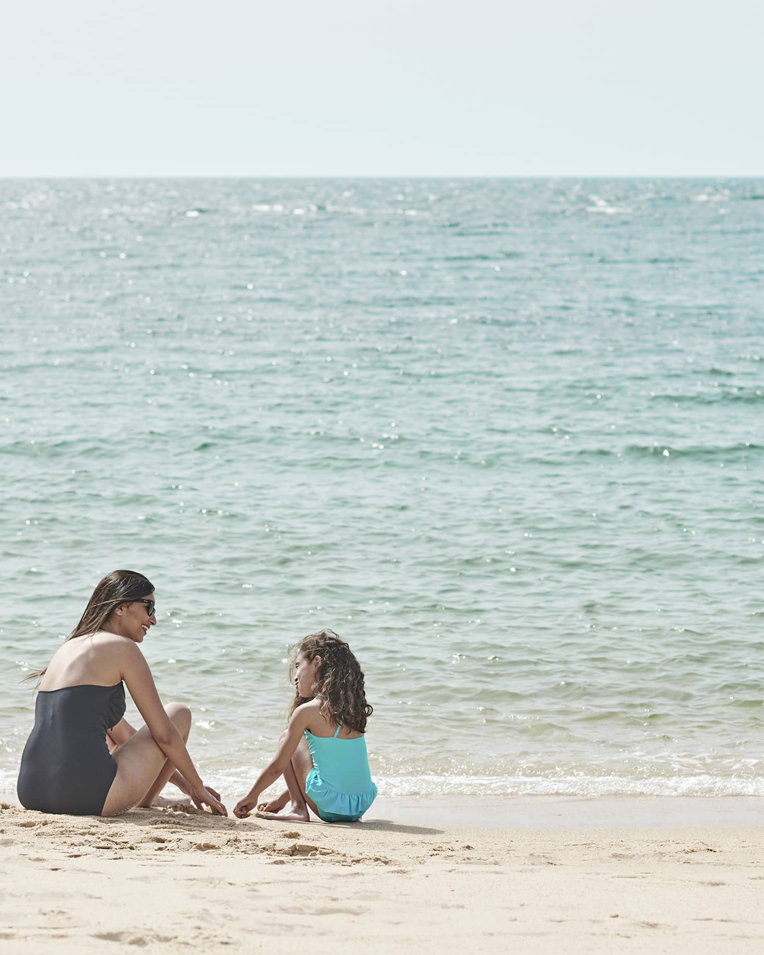 Seated by the water’s edge, a mother and daughter are having fun playing in the sand, while being kissed by the sun’s glow.