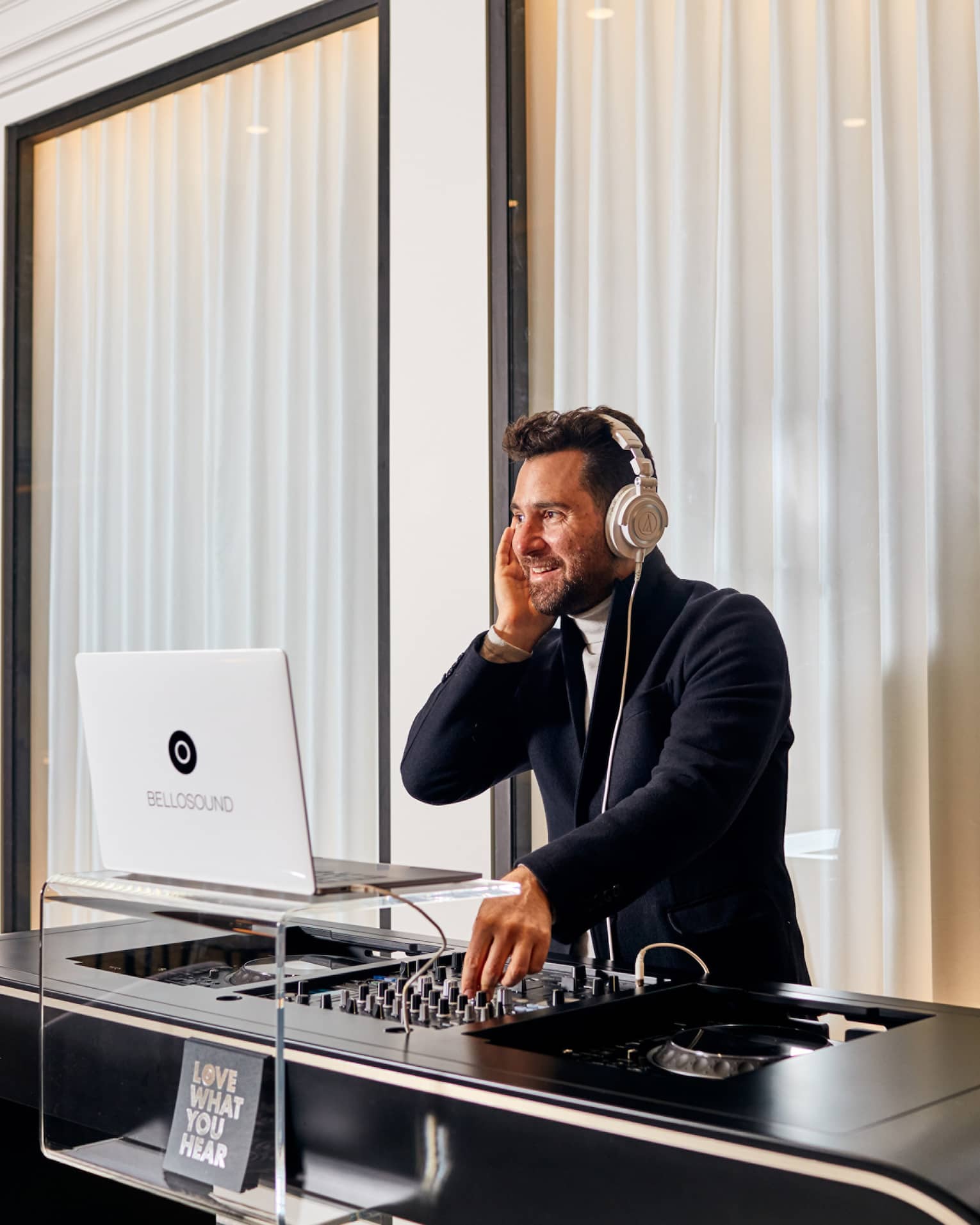 A DJ performing in a white room with closed windows behind him.