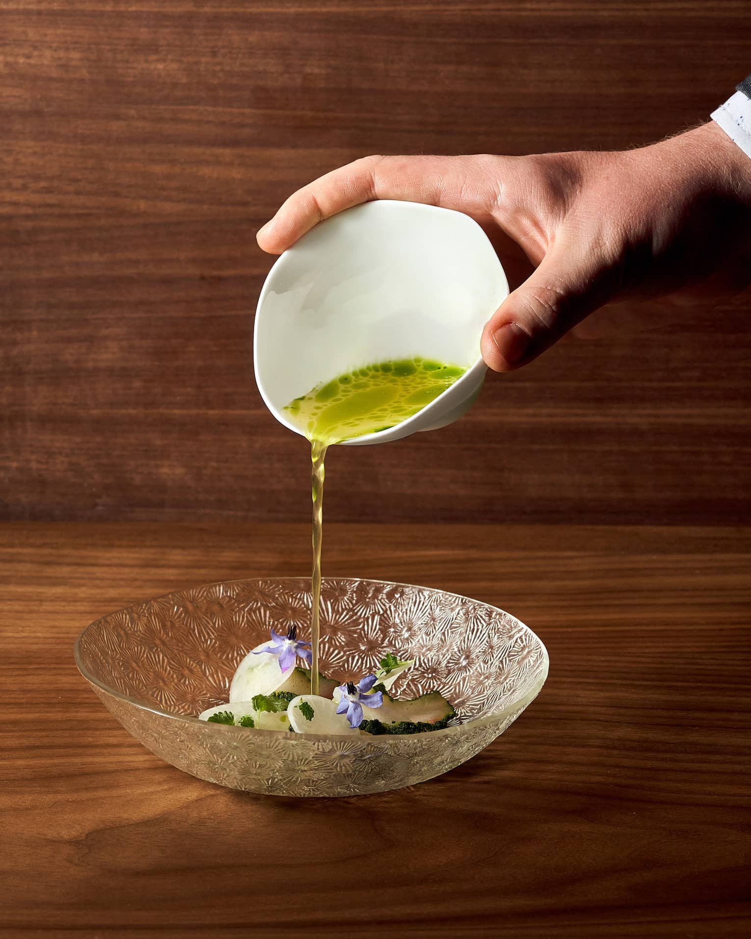 A close-up shot shows a hand delicately pouring a vibrant green sauce from a white bowl onto an intricately designed glass plate.