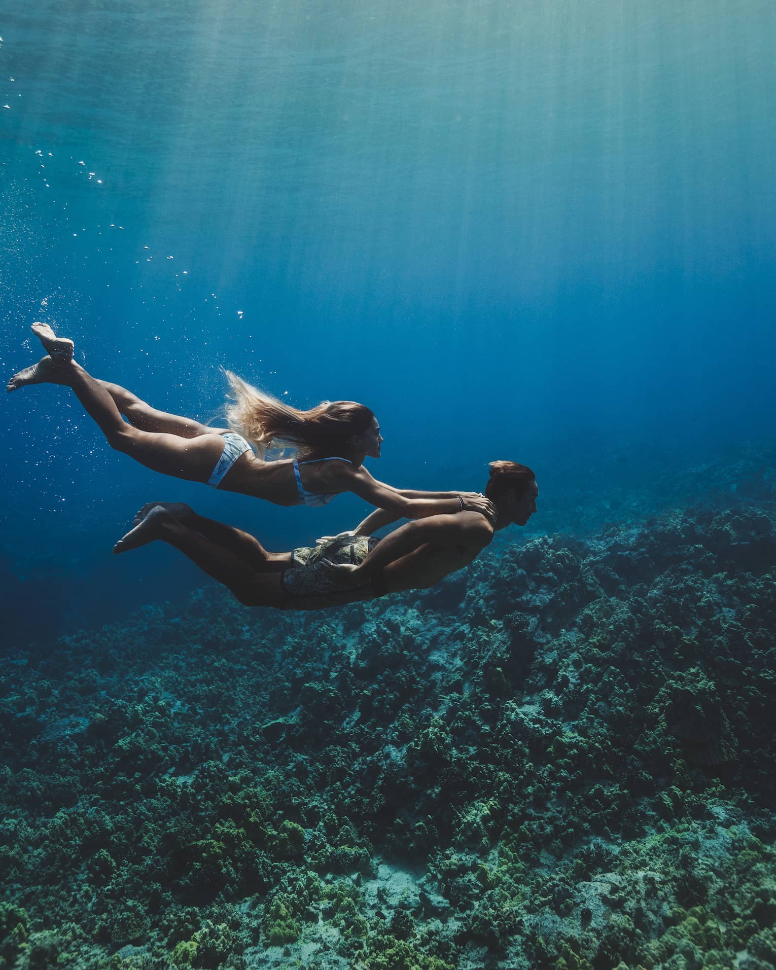 A man and woman swim underwater in the ocean over a coral reef. 