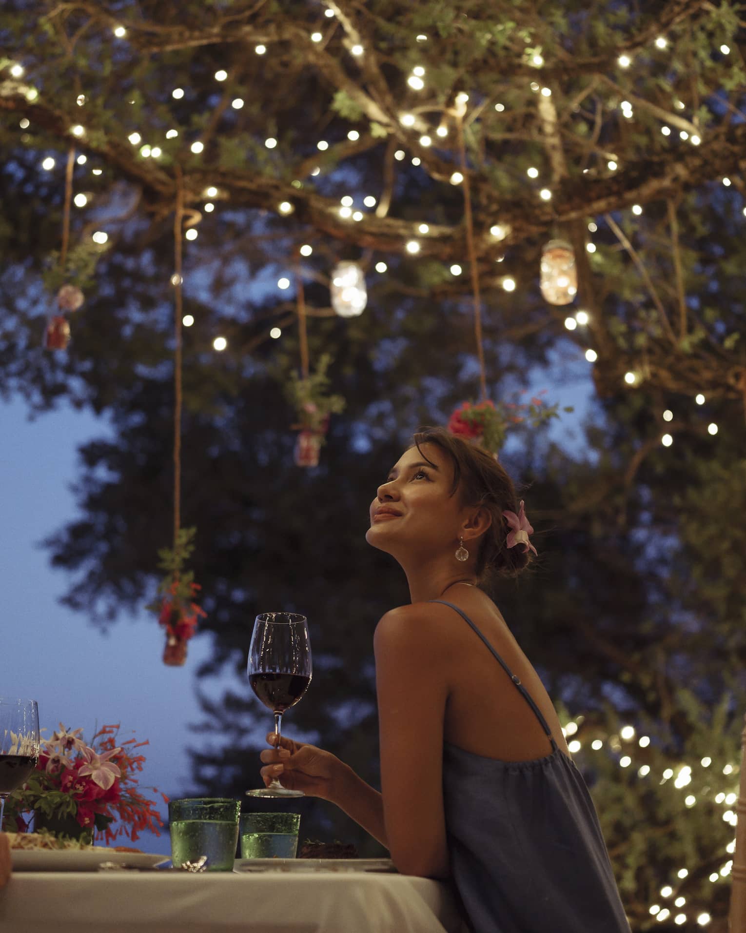 A smiling couple having a romantic dinner under trees covered in string lights and jars of flowers hanging from the branches.