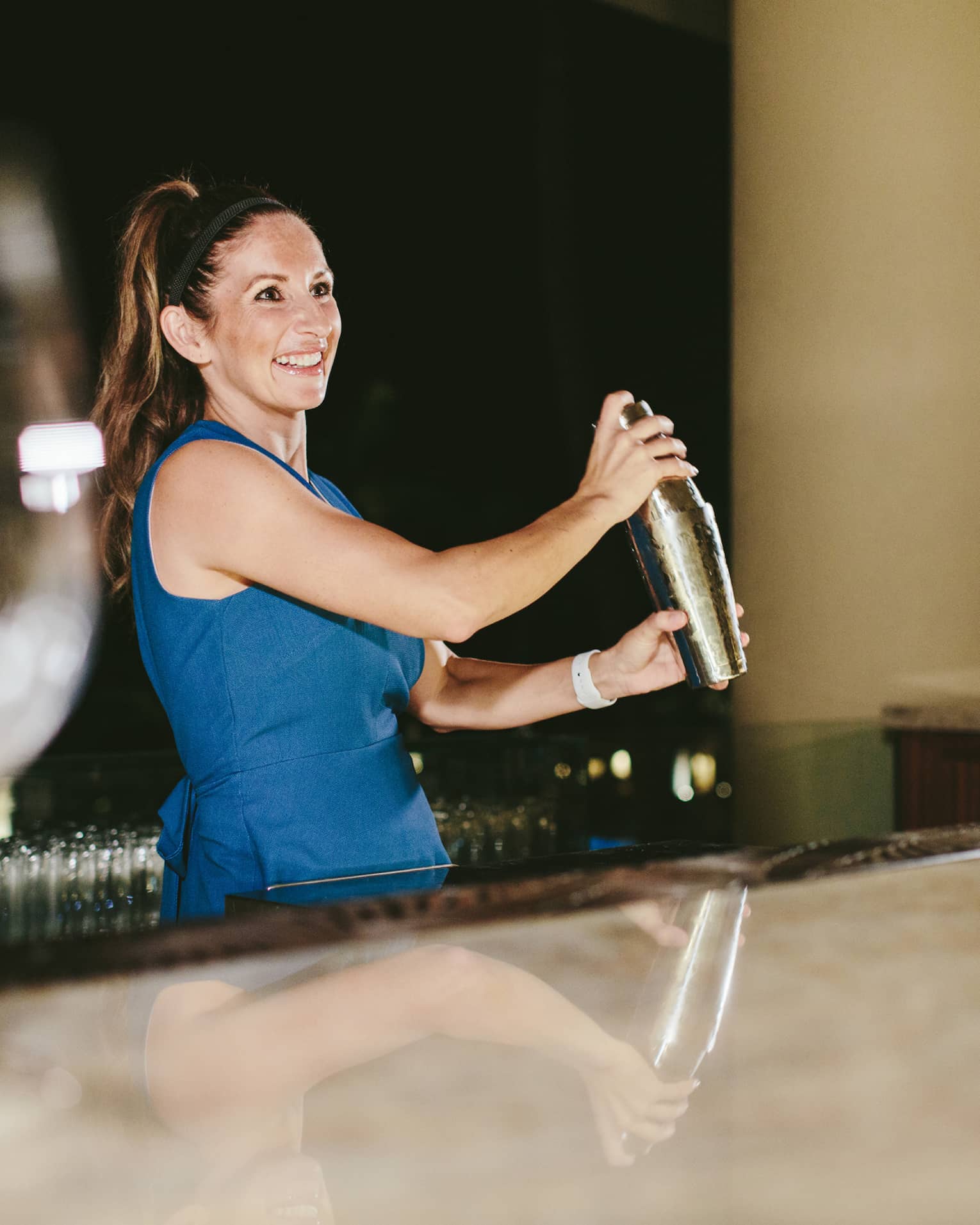 Wine goblets sparkle atop an outdoor bar in the foreground, as a smiling bartender shakes a cocktail beneath a jet-black sky.
