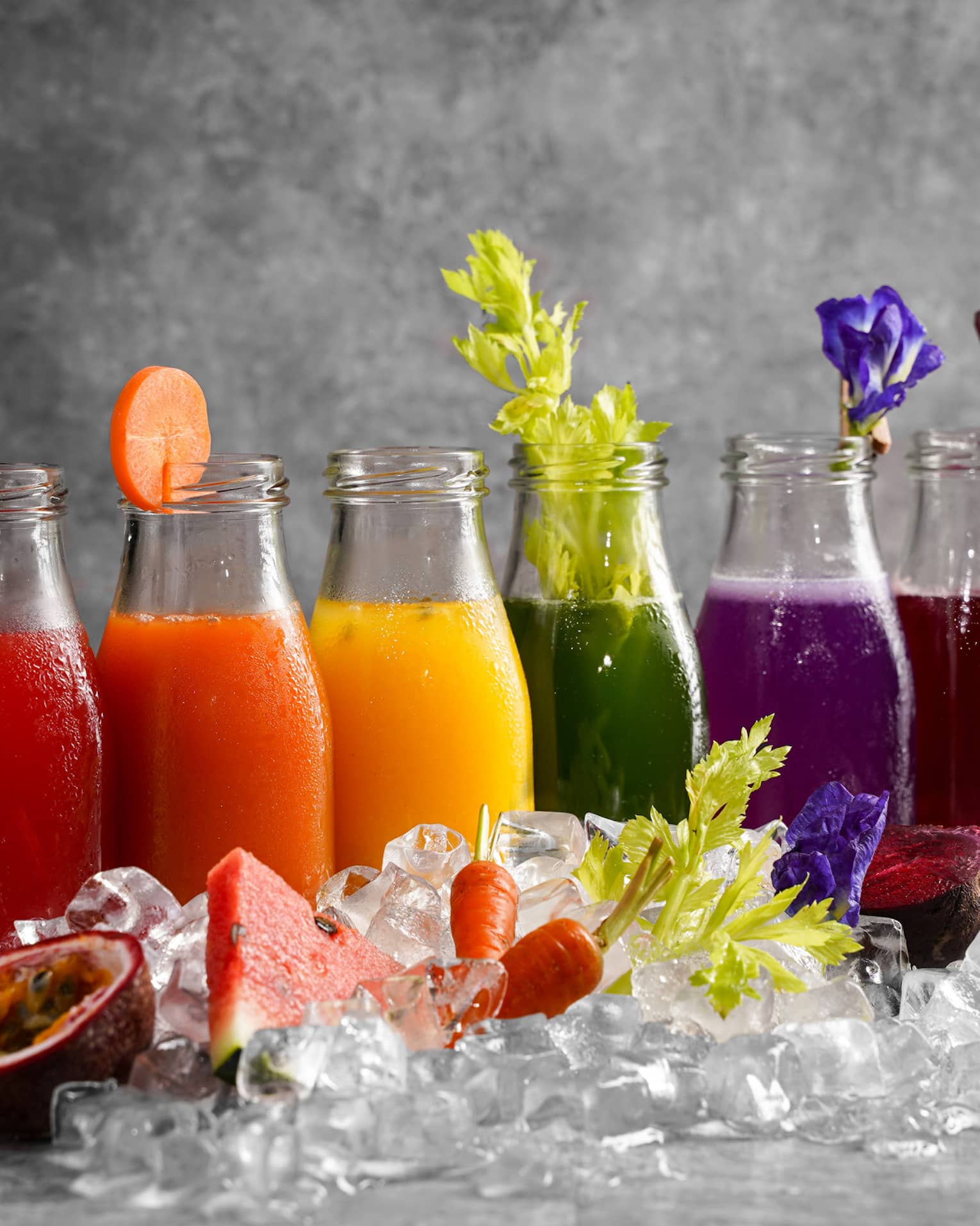 Six bottles of assorted colourful juices and garnishes lined up behind a pile of ice cubes