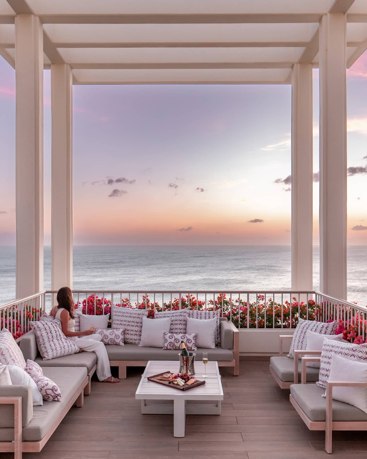 Woman sits on sofa on outdoor terrace under a white pergola overlooking the ocean