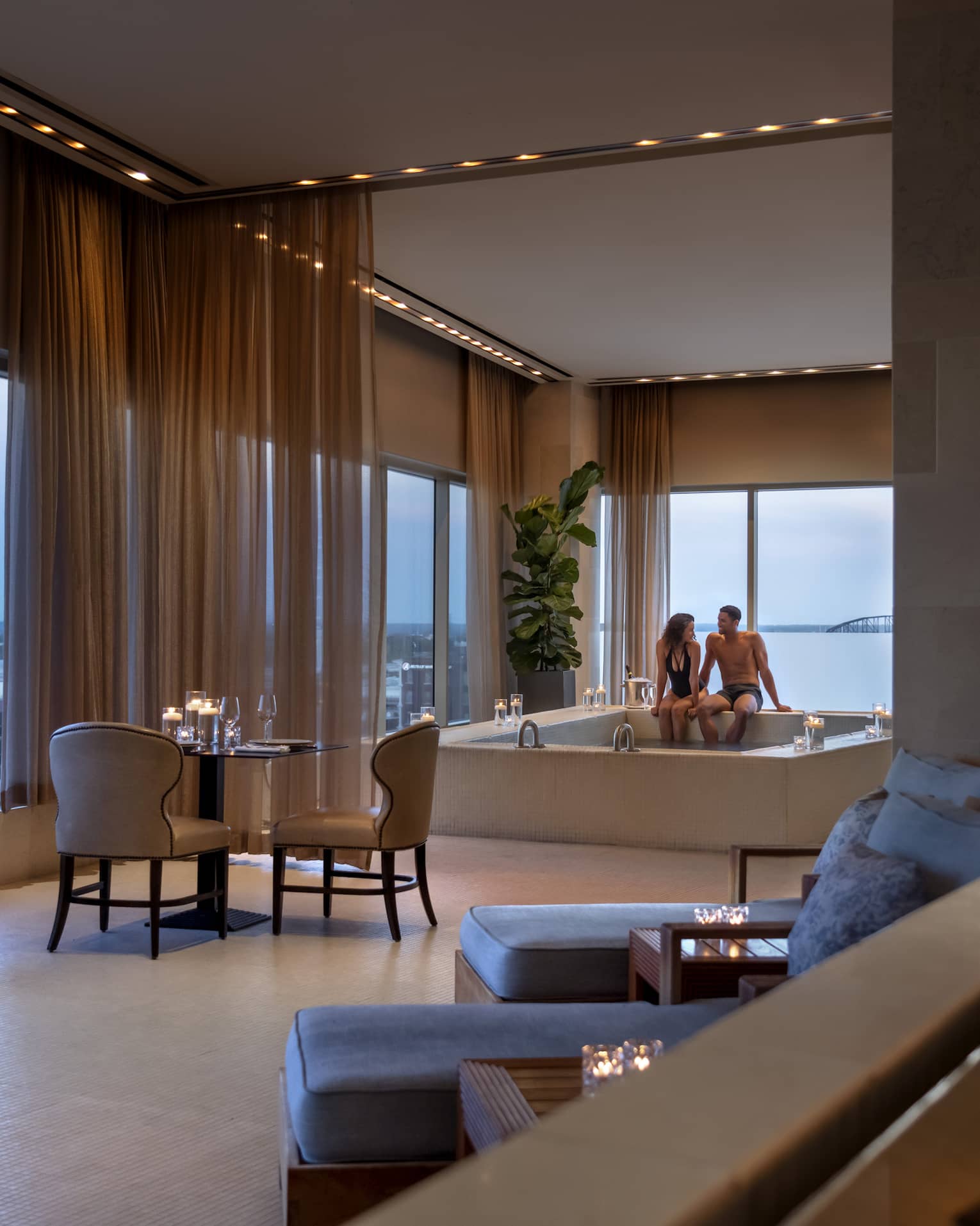 A couple enjoying a candlelit hot tub in a luxury hotel spa, with elegant seating, soft lighting and large windows offering a view of a distant bridge