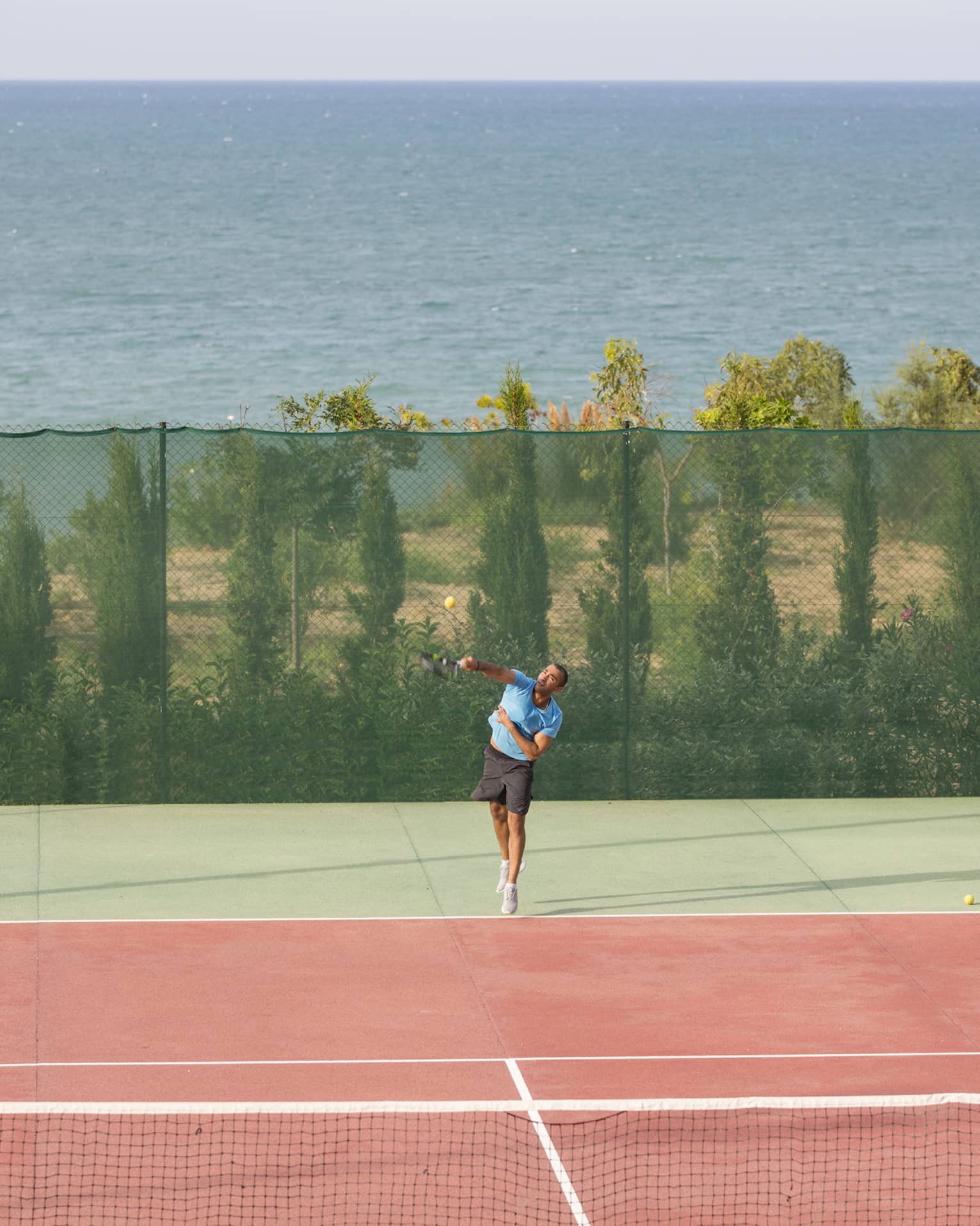 Man in blue shirt and black shorts serving a tennis ball on a seafront tennis court