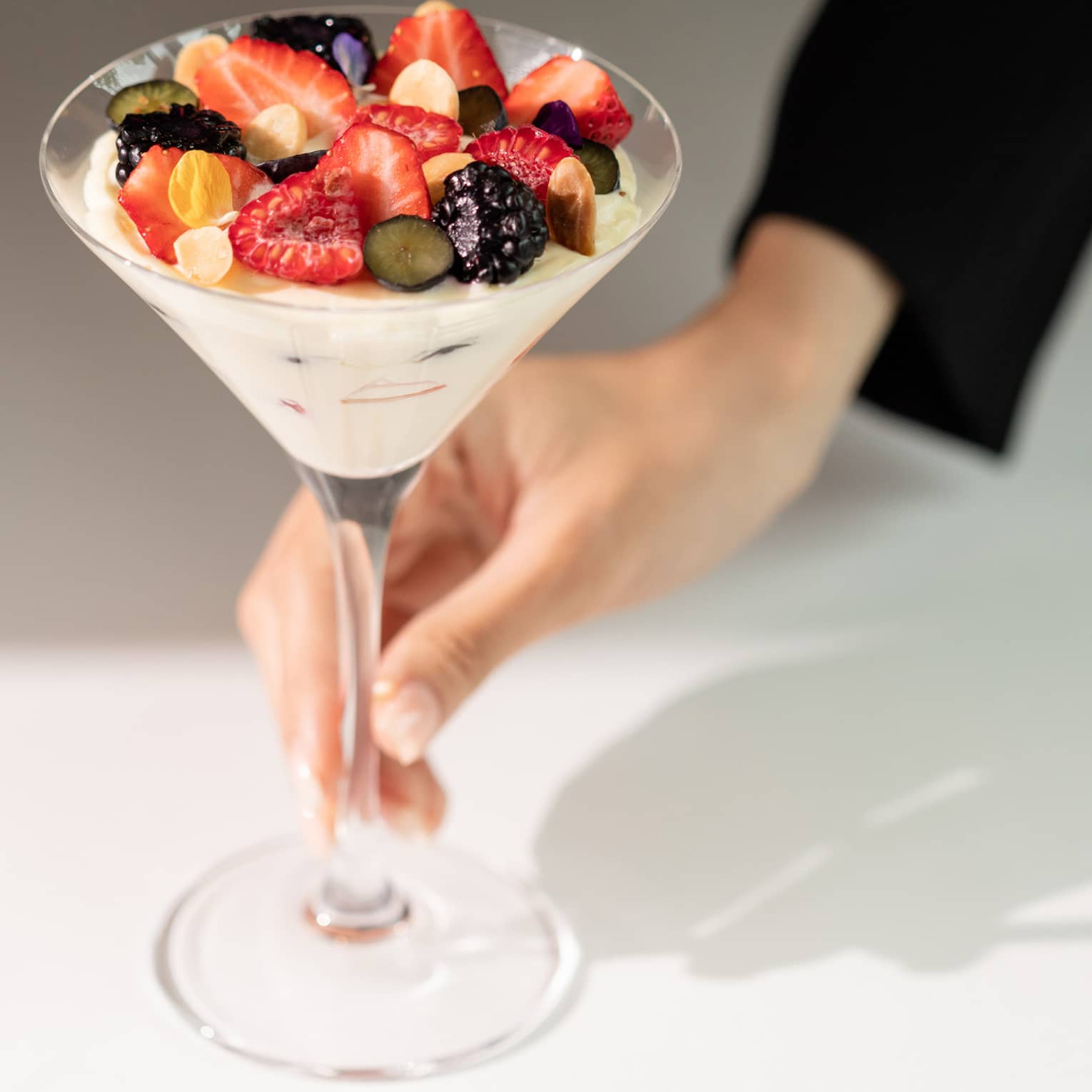 A hand delicately holds a sunlit martini glass filled with yogurt topped with summer fruits, chopped nuts, and flower petals.