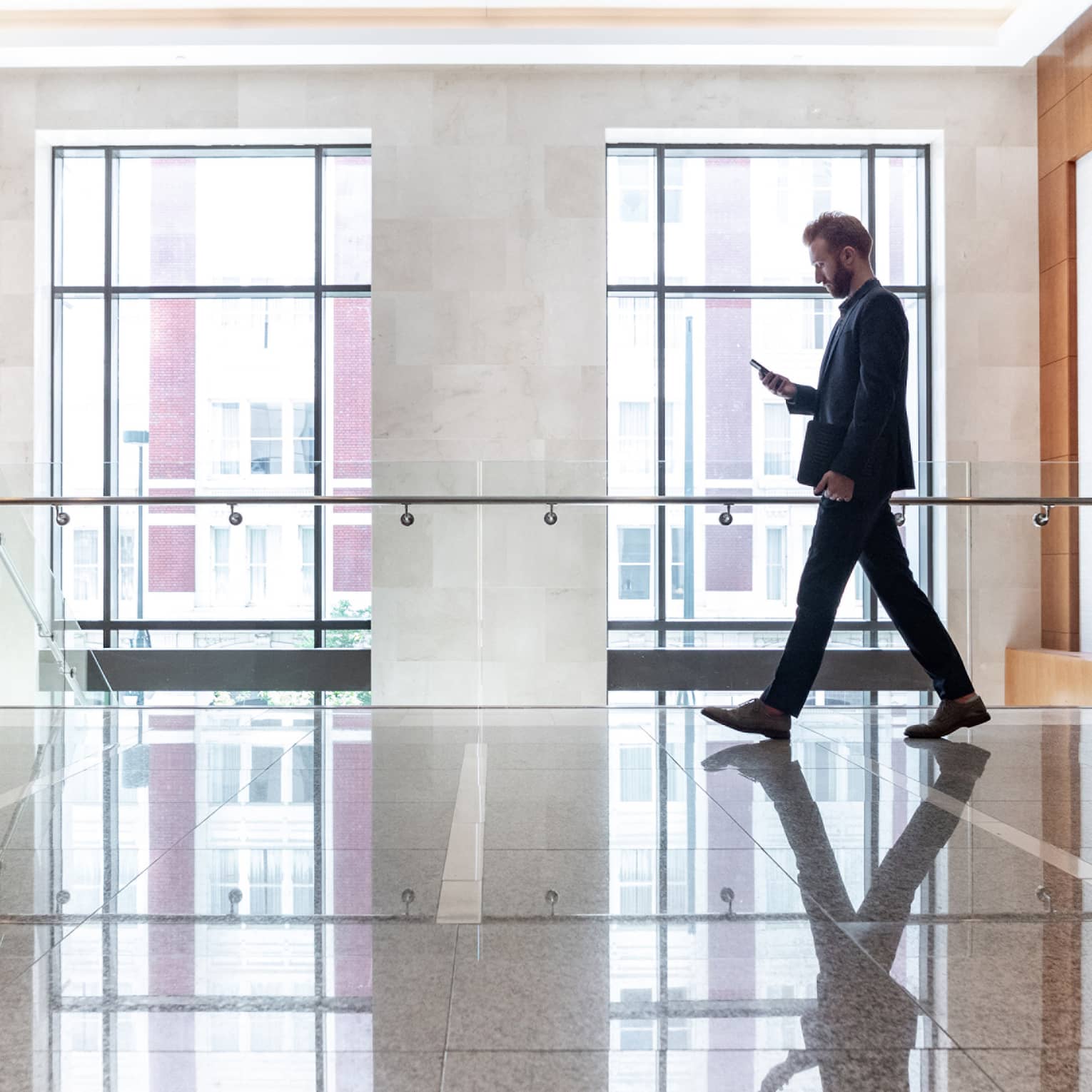 A man looking at his phone while he walks across a lobby area with large windows behind him