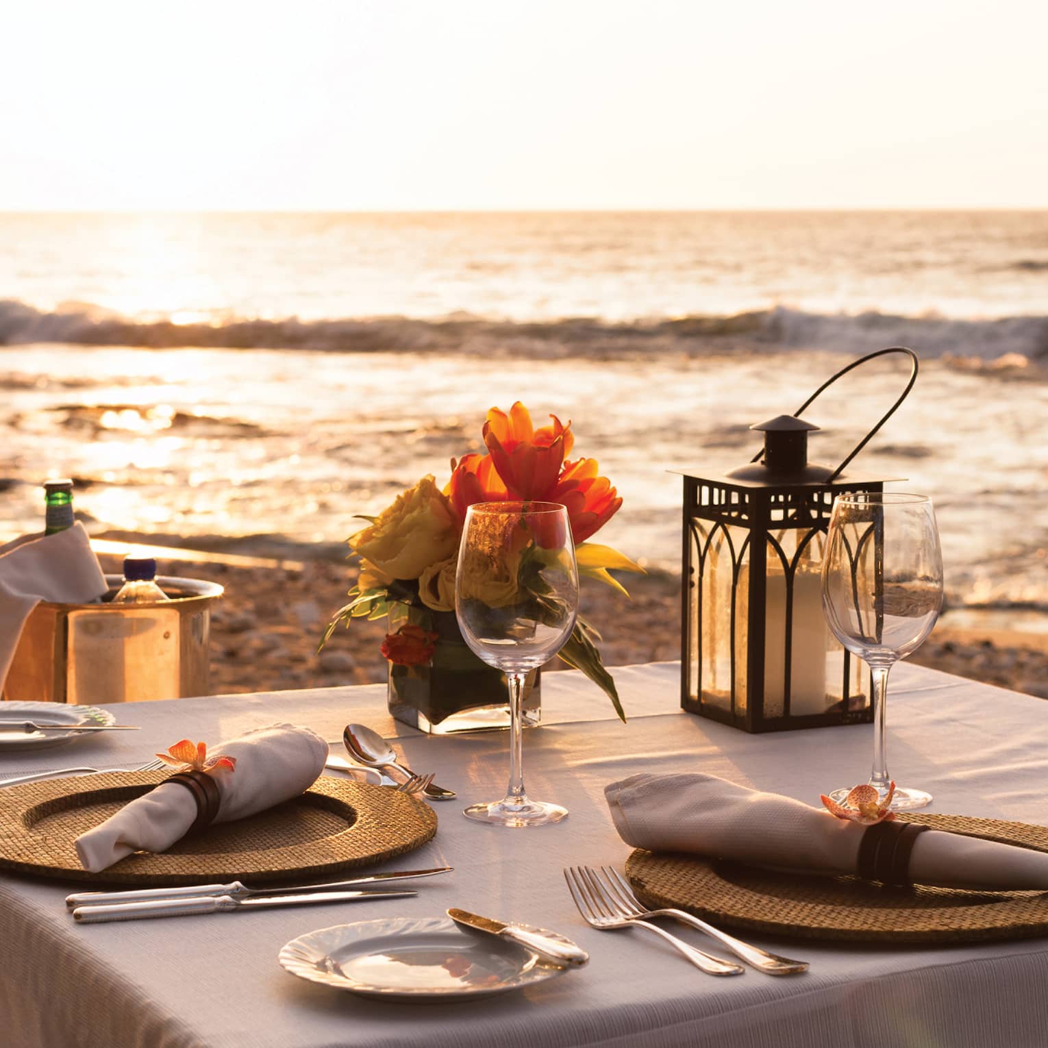 Close-up of formal place setting on table with white cloth, Champagne in bucket on beach at sunset